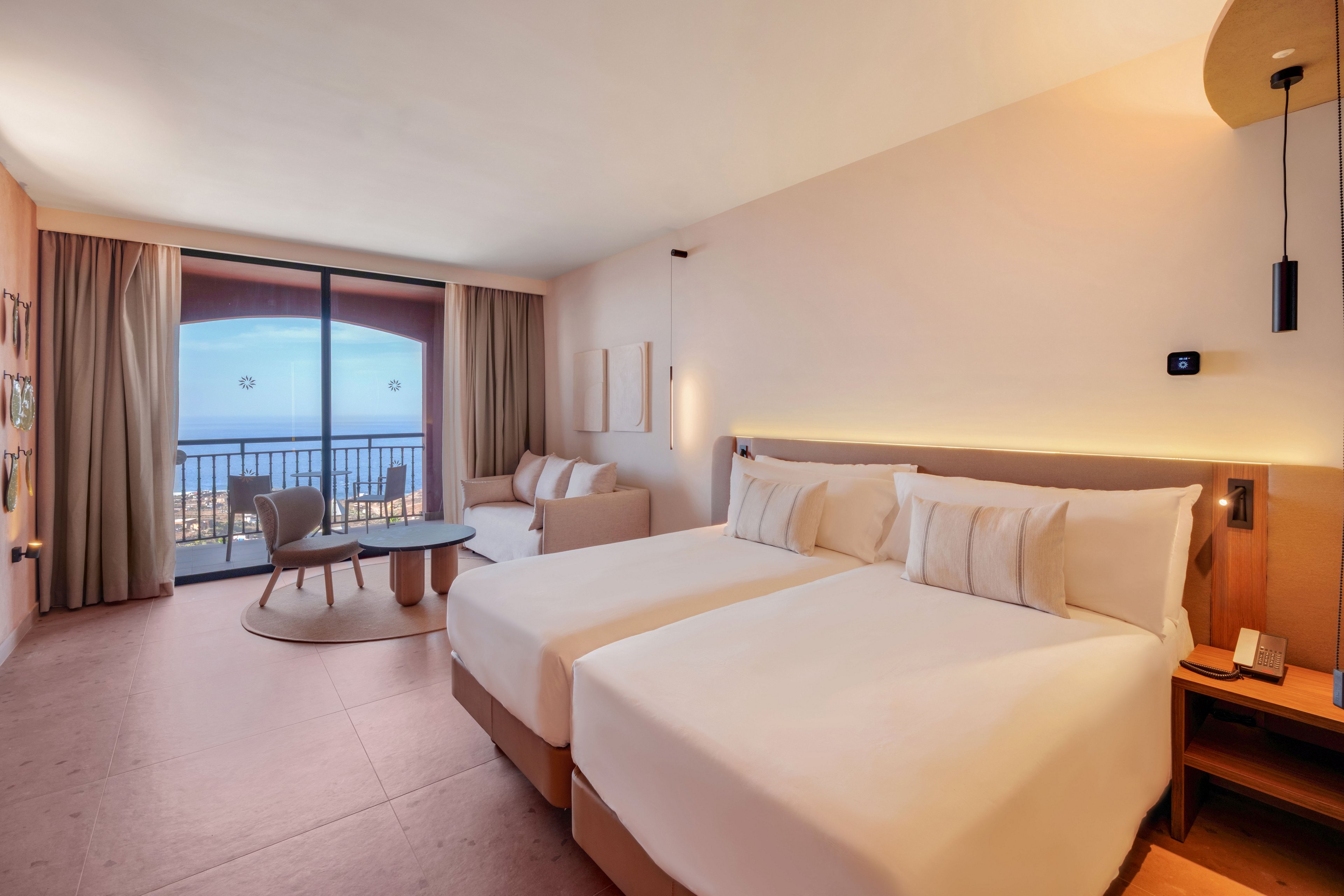 A contemporary hotel room features two neatly made twin beds with white linens and soft lighting. The space includes a small seating area with chairs and a round table near floor-to-ceiling windows. Beyond the sliding glass doors, a balcony overlooks the ocean and coastal buildings at sunset or dusk. Neutral tones and minimalist decor create a calm, relaxing atmosphere ideal for vacation or business travel.