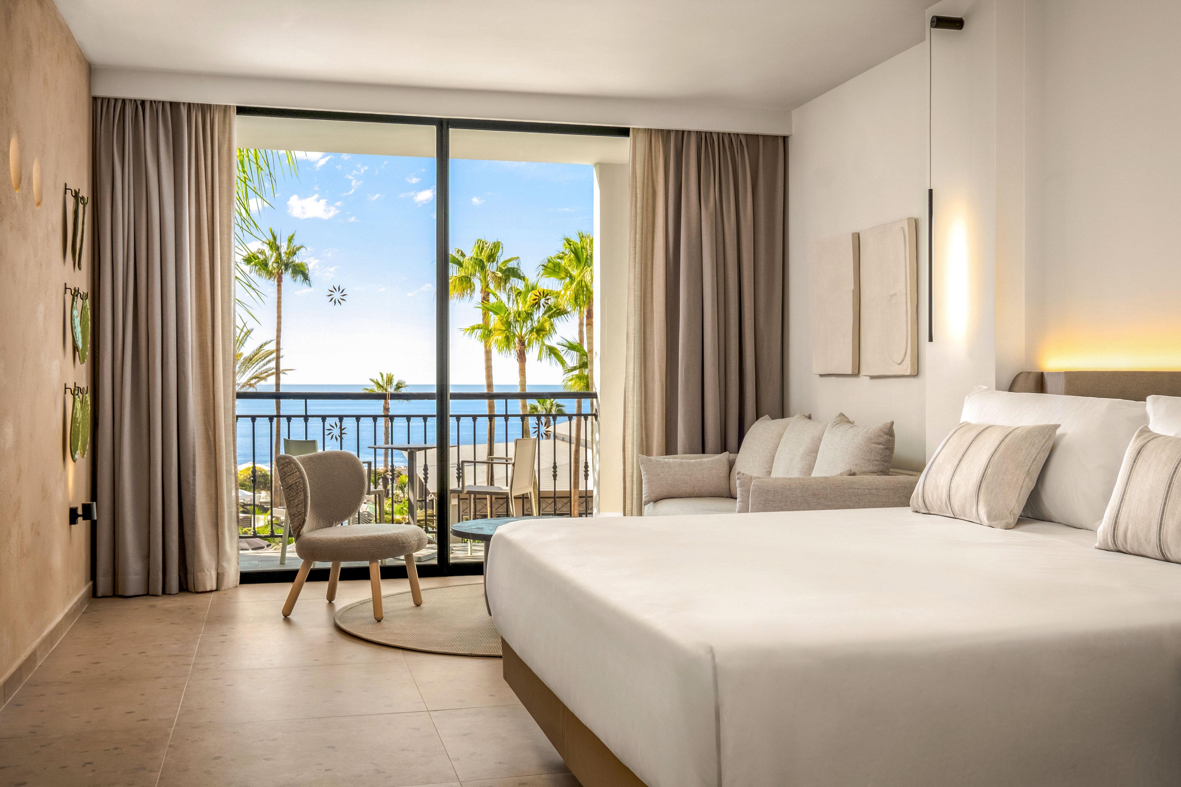 Bright contemporary hotel room features a large bed with neutral linens and cushions. A cozy seating area with a chair and sofa faces floor-to-ceiling glass doors. The balcony overlooks palm trees and a clear blue ocean under a sunny sky. Soft beige tones, minimalist decor, and natural light create a calm, relaxing atmosphere.