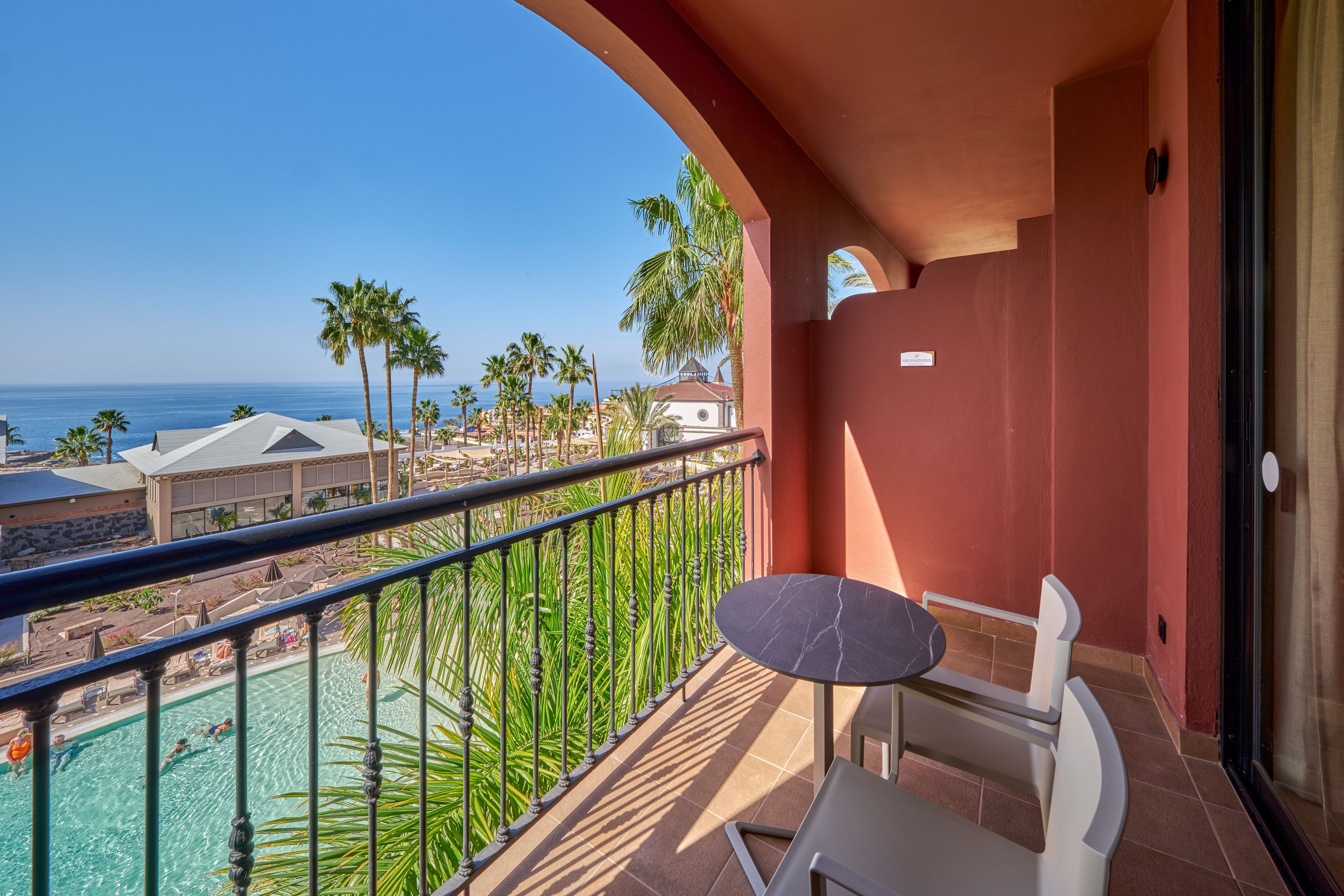 A small balcony with a round table and two chairs overlooks a resort swimming pool and the ocean beyond. Palm trees and sun loungers surround the pool area, creating a tropical vacation atmosphere. The building features warm terracotta walls and an arched opening framing the scenic view. Clear blue sky and calm water suggest a sunny, relaxing day at a coastal hotel.