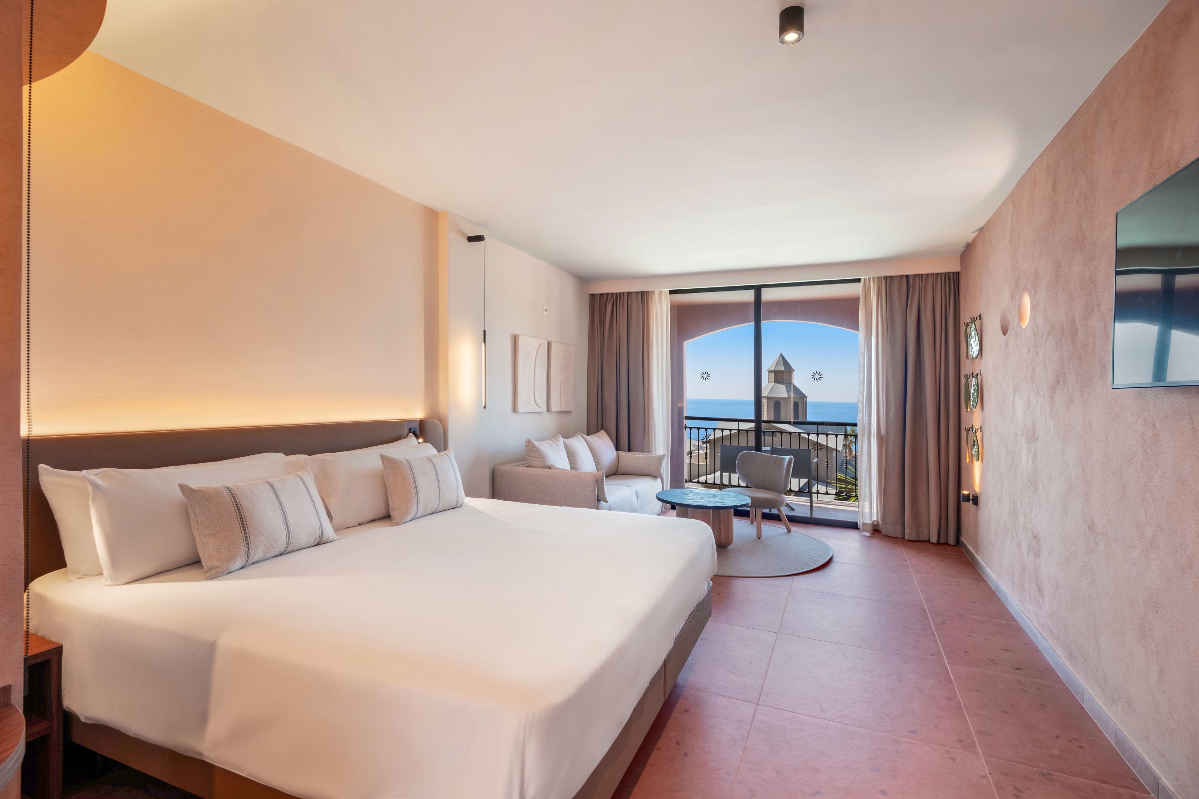 A spacious modern hotel room features a large bed with multiple pillows and soft neutral tones. The room includes a seating area with a sofa and round coffee table near floor-to-ceiling windows. Curtains frame a balcony that overlooks a scenic coastal view with a tower and the ocean in the distance. Warm lighting and minimalist decor create a calm, inviting atmosphere.