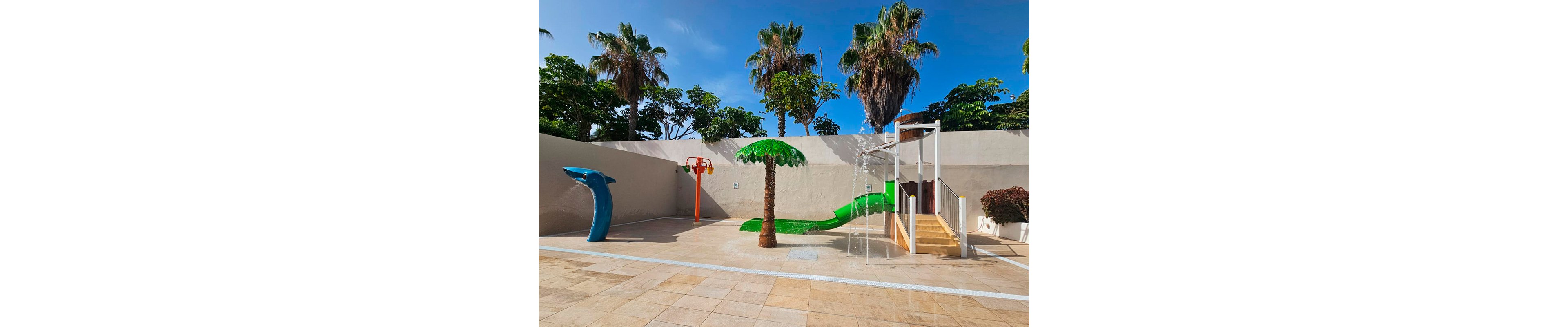 An outdoor children’s splash zone features playful water structures under a bright blue sky. A green slide and palm tree fountain stand on a wet play surface beside a small stair-and-platform structure with water jets. A dolphin-shaped sprayer and orange overhead shower add to the fun aquatic theme. Tall palm trees and a high wall enclose the sunny recreation area, creating a safe and vibrant space for kids.