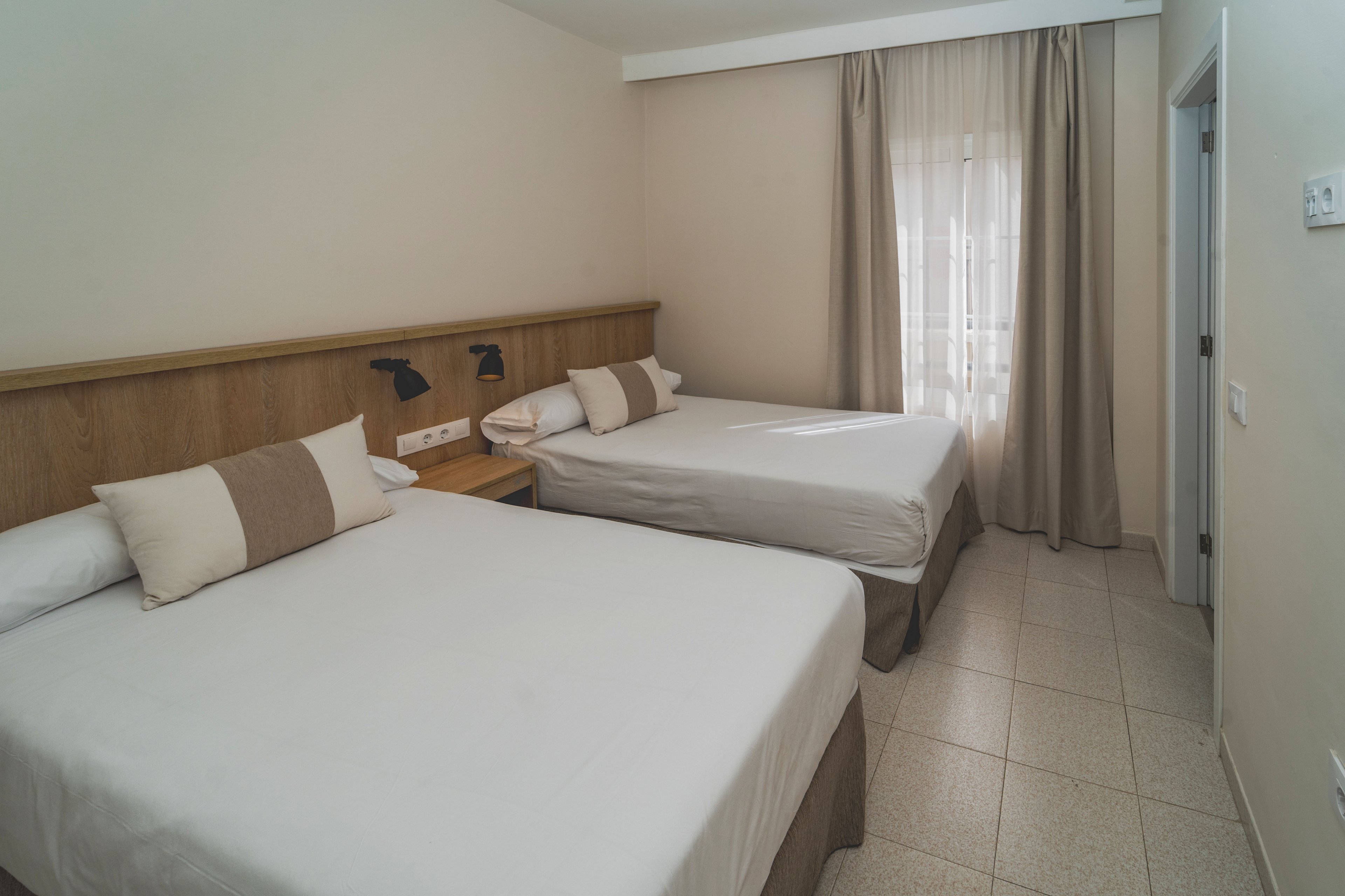 A simple hotel room features two neatly made double beds with white linens and beige accent pillows. The space has light-colored tiled flooring and a wooden headboard with mounted reading lights. Natural light enters through a window covered with sheer and beige curtains, creating a soft, inviting atmosphere. The overall design is minimal, clean, and functional, suitable for travelers or guests.
