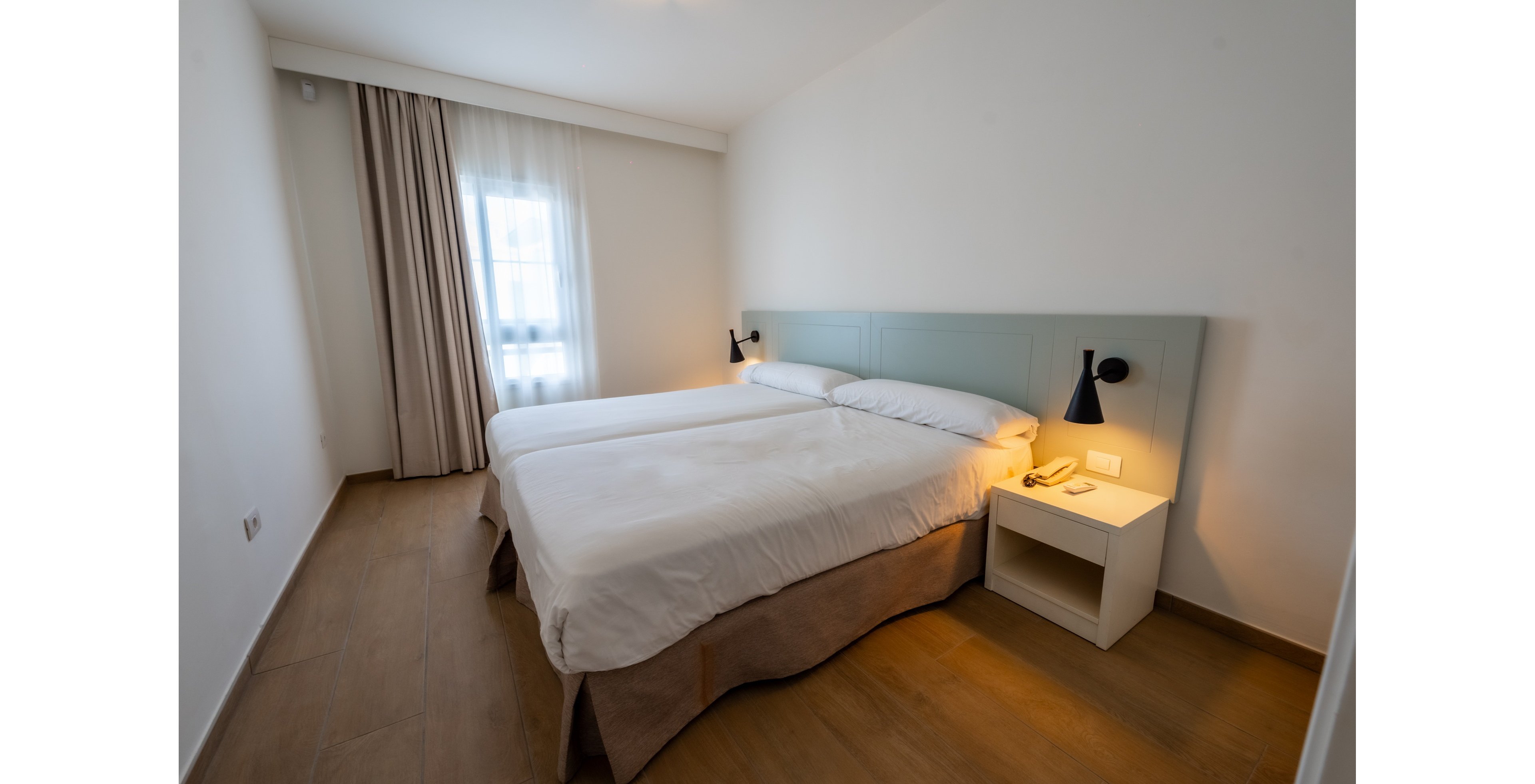 A simple, modern bedroom features two twin beds pushed together with white linens and a light-colored headboard. The room has wooden flooring, white walls, and a single bedside table with a telephone and wall-mounted reading lamp. Natural light enters through a window covered by sheer curtains and heavier drapes, creating a soft, cozy atmosphere. The overall design is clean, uncluttered, and contemporary.