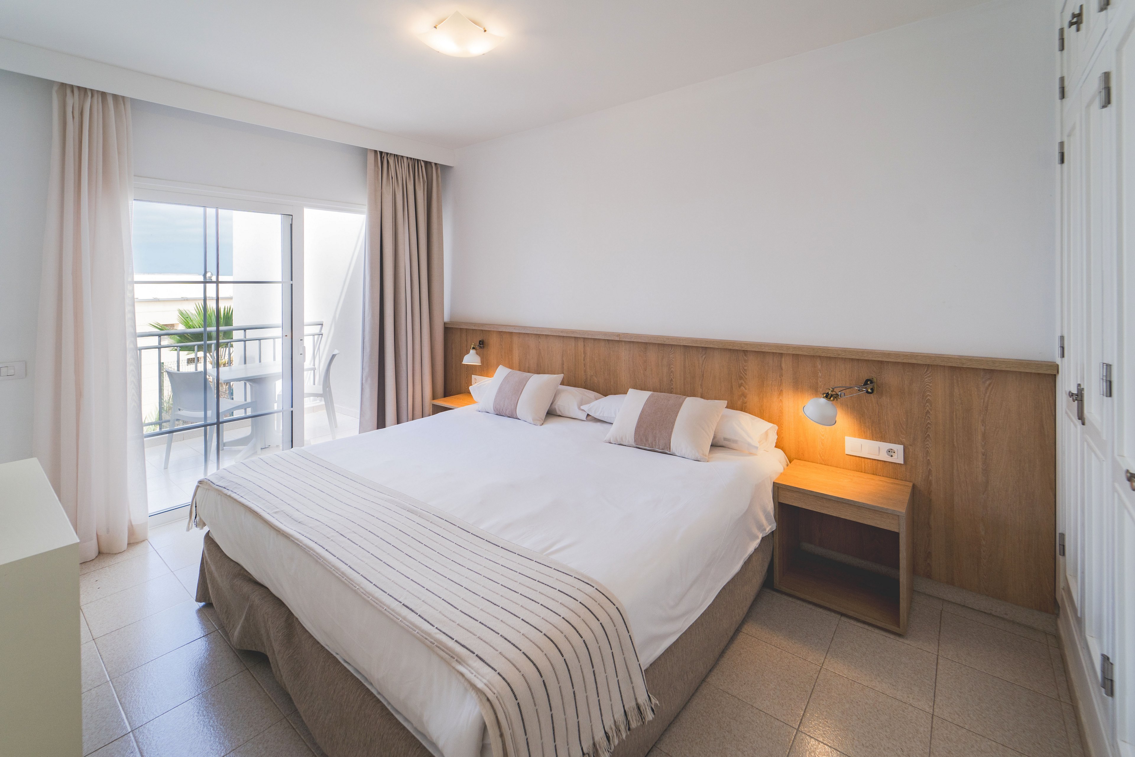 A spacious, modern bedroom features a large double bed with white linens and neutral striped throw. Wooden bedside tables with wall-mounted reading lamps flank the bed, adding a warm accent to the minimalist decor. Floor-to-ceiling curtains frame a glass door that opens onto a balcony with outdoor seating and a partial sea view. The room is clean, bright, and inviting, with soft natural light enhancing the calm atmosphere.