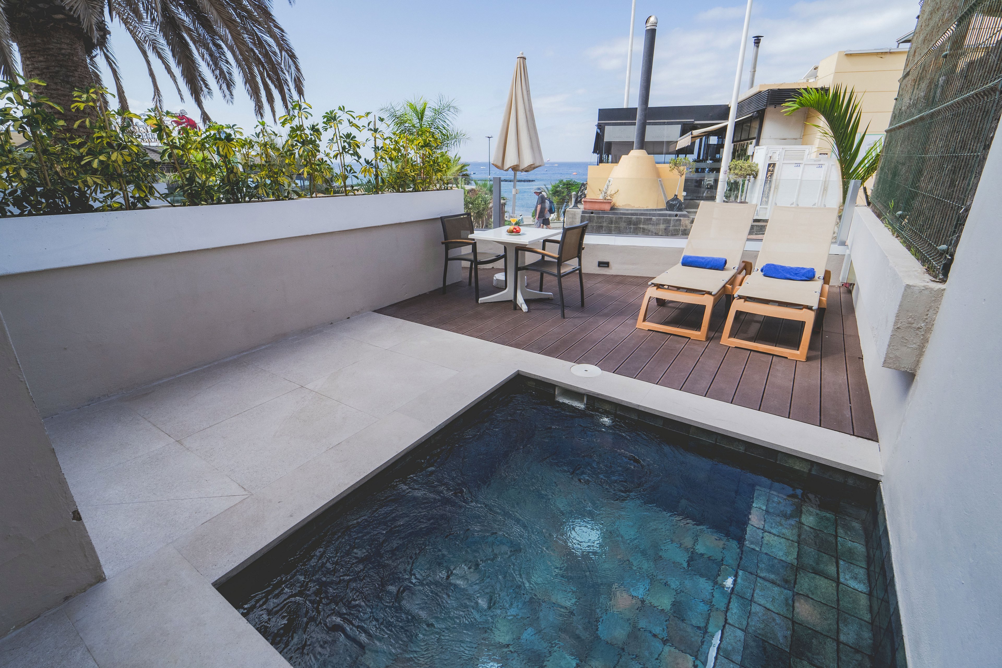 A small private terrace features a square plunge pool with tiled interior in the foreground. Beside the pool, a wooden deck holds two sun loungers with blue cushions and a round table with chairs and a closed umbrella. Green plants line the white walls, and neighboring buildings and distant sea views appear in the background. The scene suggests a relaxed vacation setting with contemporary outdoor design.