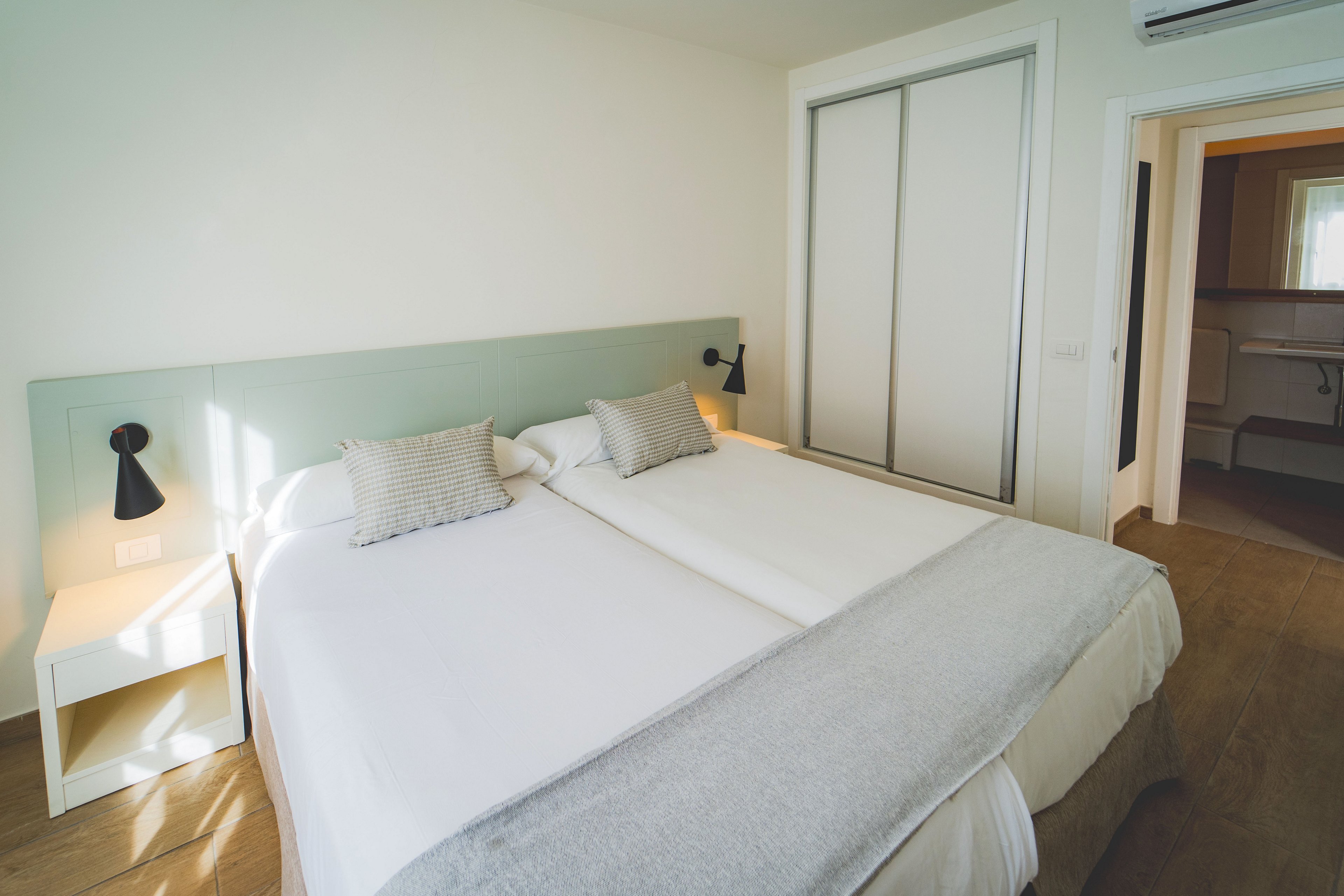 A contemporary bedroom features two neatly made twin beds pushed together with white linens and light gray throws. Minimalist bedside tables and black wall-mounted lamps frame the headboard. A large sliding-door wardrobe is visible against the wall, and an open doorway leads to an adjoining room with natural light. The space has a clean, airy feel with light wood flooring and soft neutral colors.