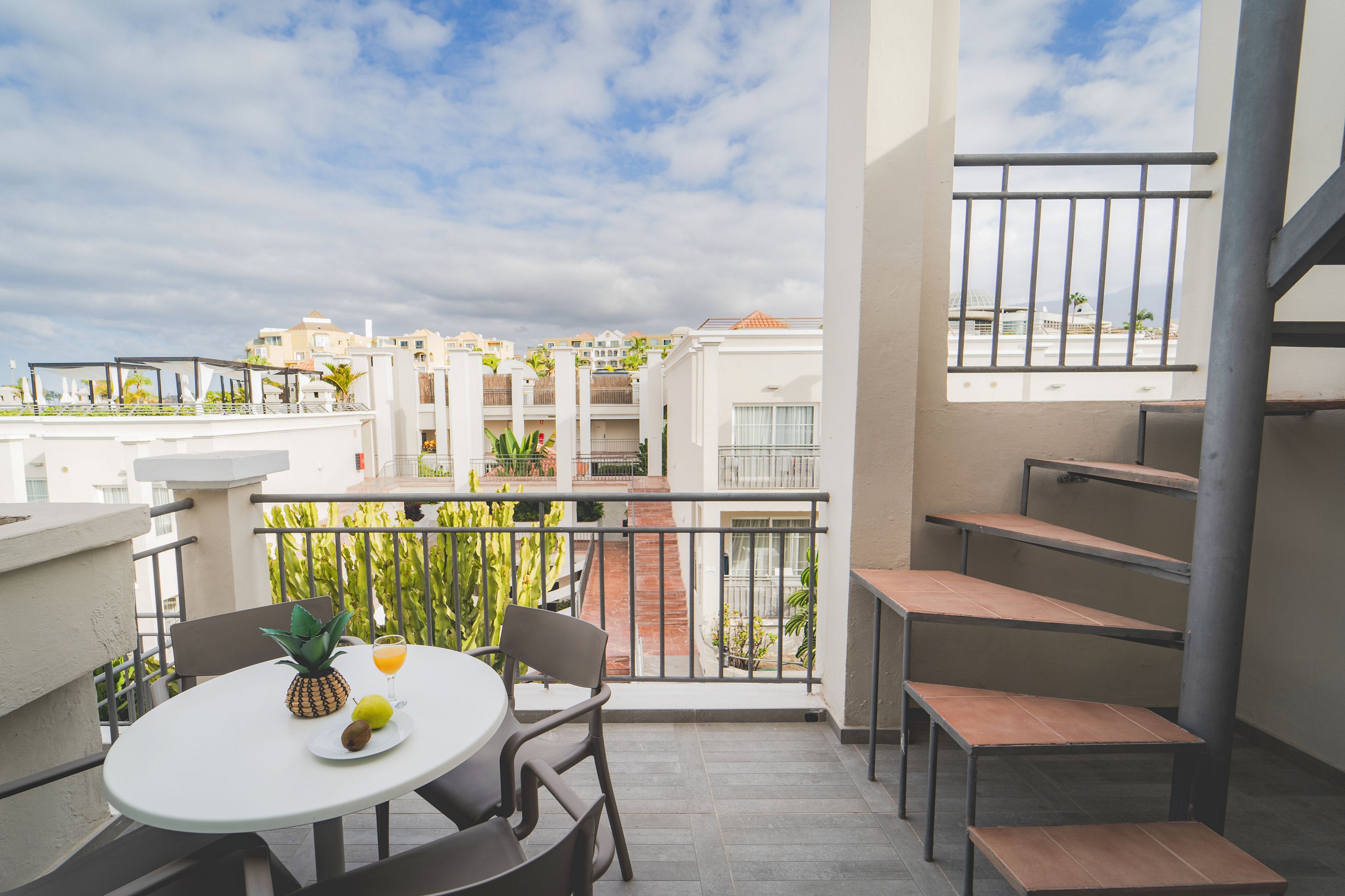 A bright apartment terrace features a round white table with several chairs and fresh fruit arranged in the center. The balcony overlooks a residential complex of white buildings under a partly cloudy sky. A modern metal and wood staircase rises beside the seating area, adding architectural interest. Green plants and clean lines create a relaxed, contemporary vacation atmosphere.