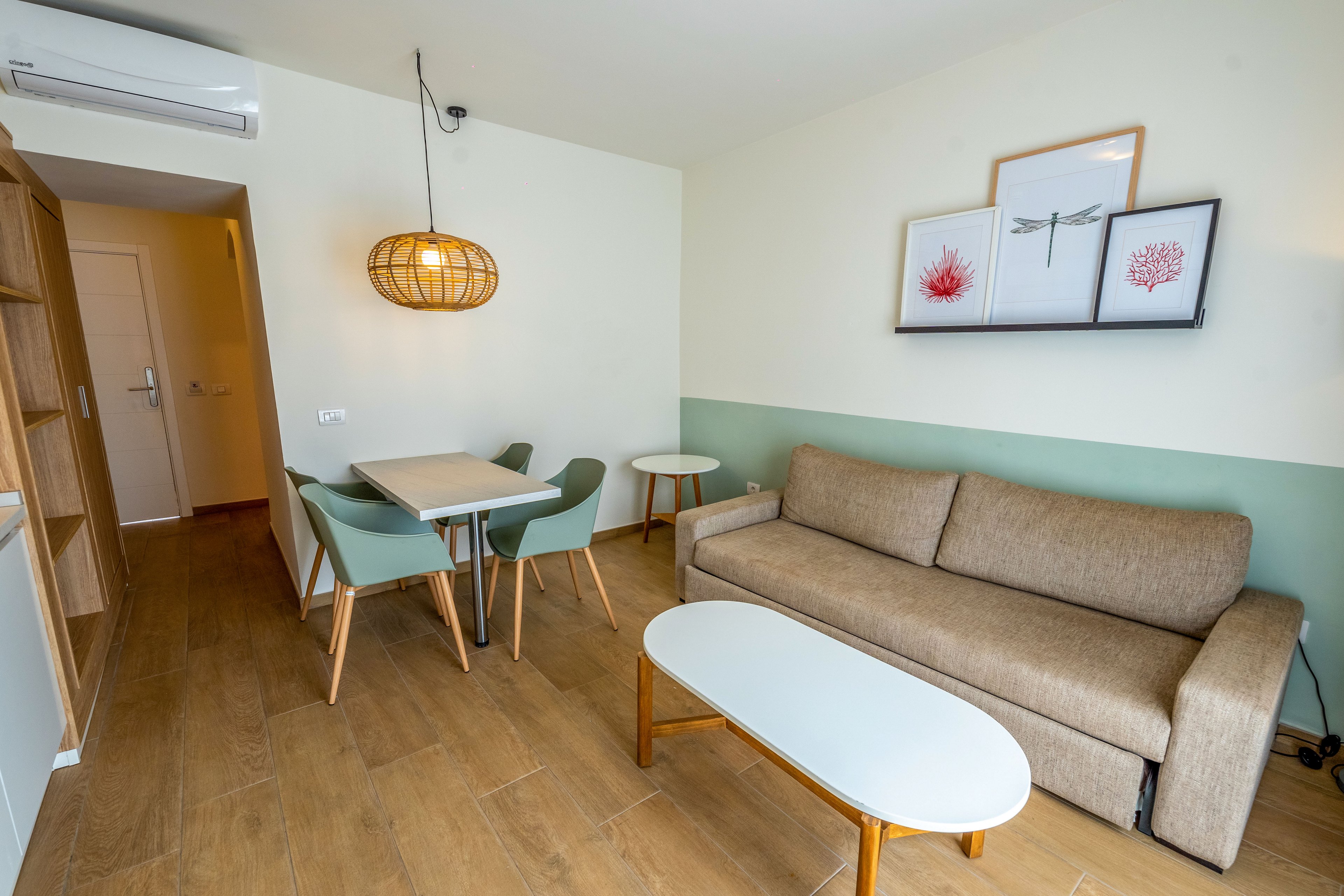 Bright contemporary living room features a beige sofa and oval coffee table on wood-look flooring. A small dining area with light green chairs and a rectangular table sits beneath a woven pendant light. Minimalist wall art with coral illustrations decorates the space above the sofa. The room includes a doorway leading to a hall and a visible air conditioning unit mounted near the ceiling.