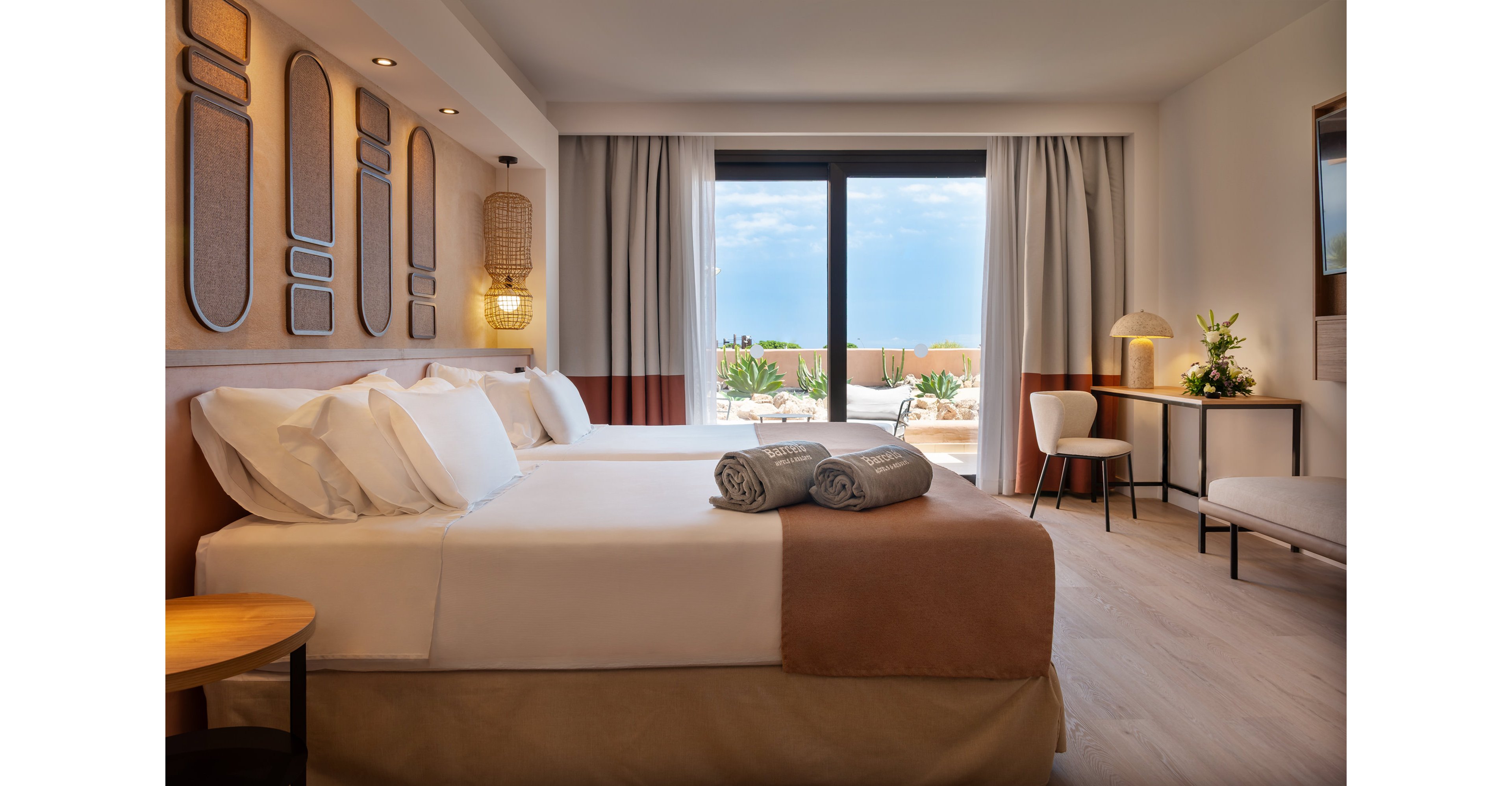 A contemporary hotel room features two neatly made twin beds with white linens and folded gray towels placed on top. The space is decorated in warm neutral tones, with textured wall panels, soft lighting, and a minimalist desk and chair. Large glass doors open to an outdoor terrace with plants and a view of a bright blue sky. The overall atmosphere is calm, inviting, and designed for comfortable vacation or business stays.