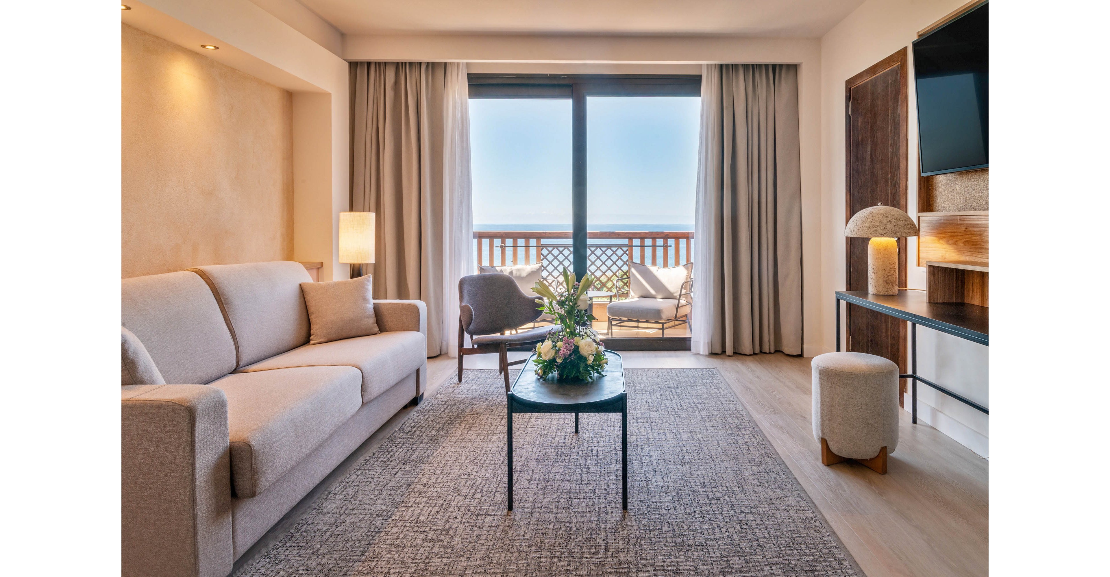 A contemporary hotel suite features a light beige sofa and a central coffee table with a fresh floral arrangement. Floor-to-ceiling windows with curtains open onto a balcony overlooking a bright blue sea. A lounge chair and small table sit outside, while a modern console, television, and upholstered stool complete the interior. Soft neutral tones, warm lighting, and clean lines create a calm, inviting atmosphere.