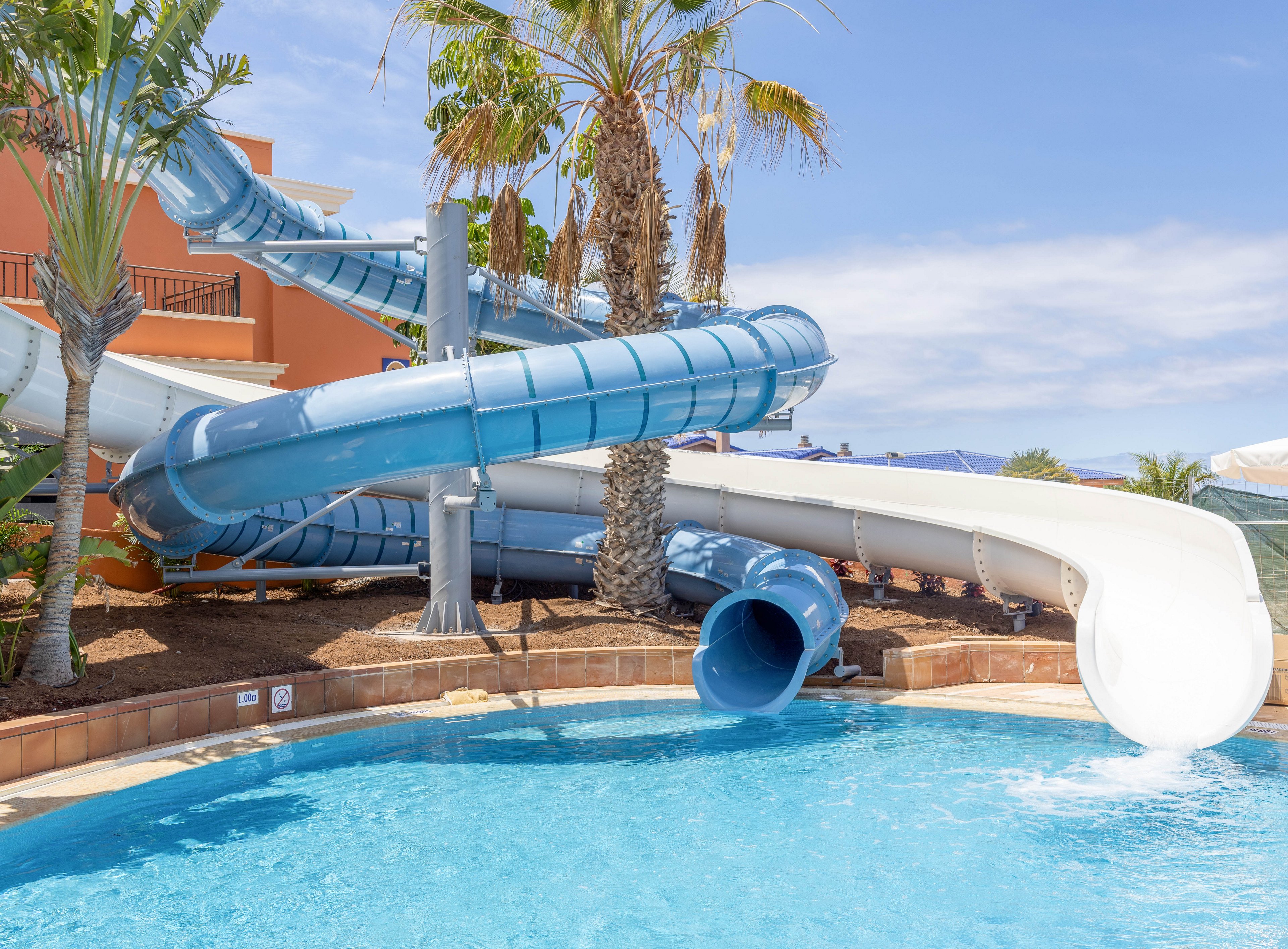 A bright blue swimming pool sits at the base of several twisting waterslides in an outdoor resort setting. The slides curve around palm trees and landscaped areas before emptying into the clear water. Warm sunlight and a clear sky create a summery, vacation atmosphere. Nearby buildings and shade structures suggest a family-friendly water park or hotel pool area.
