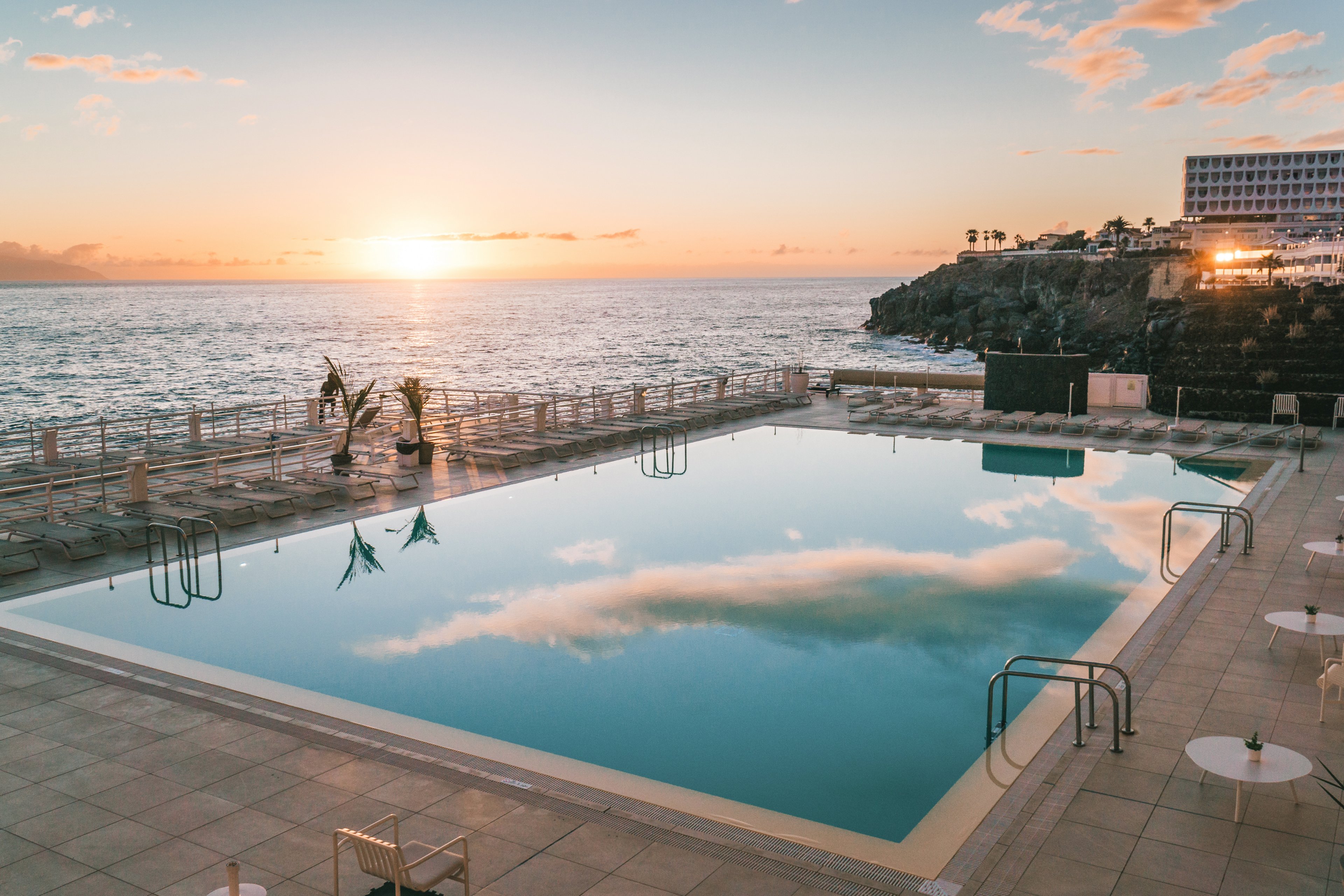 A large, empty swimming pool reflects soft clouds and the colorful sky at sunset. The pool is situated on a spacious tiled terrace overlooking the open ocean. Metal ladders, railings, and a few scattered tables and chairs frame the serene scene. In the distance, a rocky coastline and a modern hotel building rise above the water, adding depth to the coastal landscape.