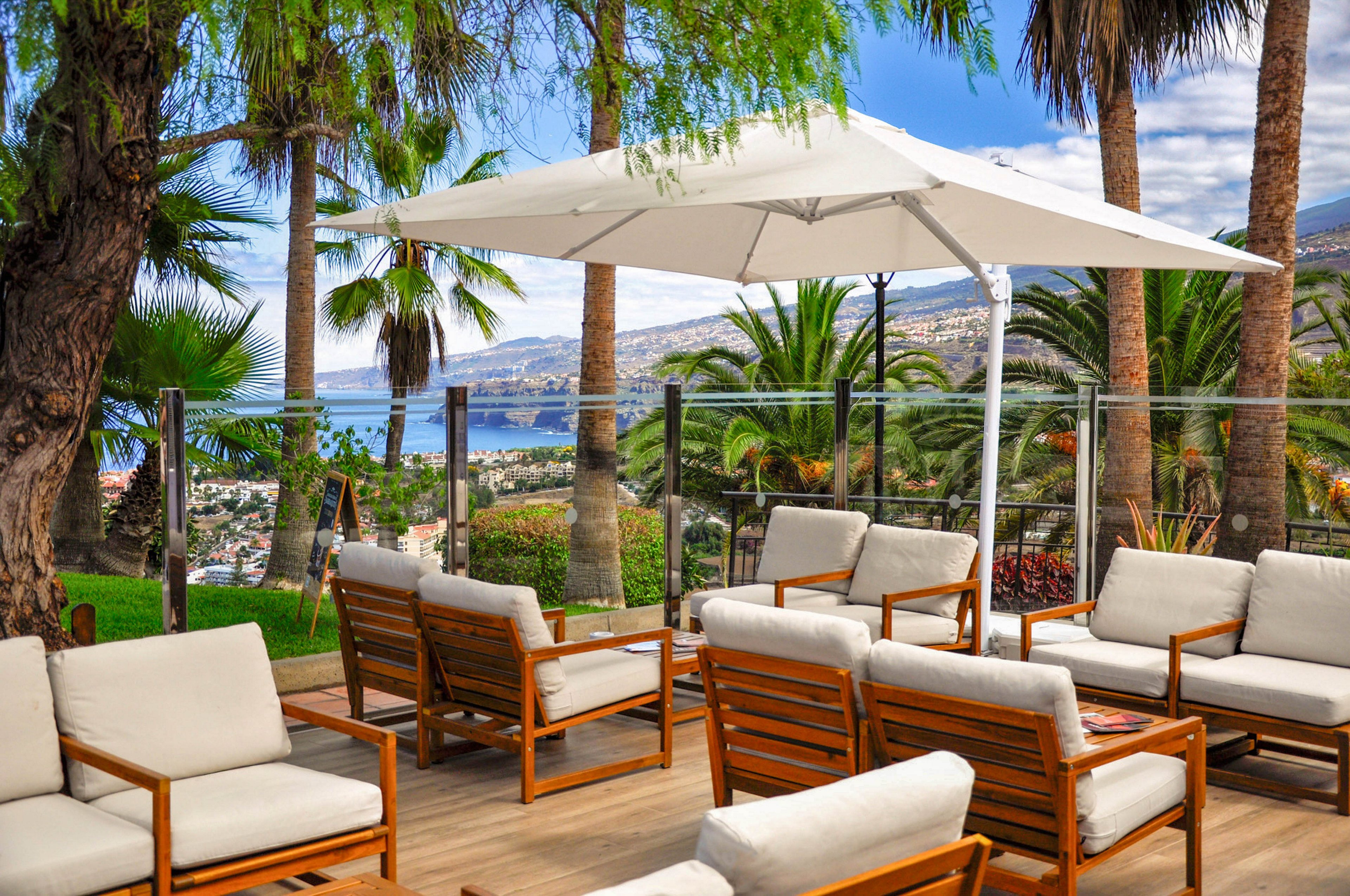 Outdoor lounge chairs with white cushions sit on a wooden terrace under a large white umbrella. Palm trees and lush greenery surround the seating area, creating a tropical atmosphere. Glass railings frame a scenic view of a coastal town and blue ocean in the background. The bright daylight and clear sky suggest a relaxing vacation or resort setting.