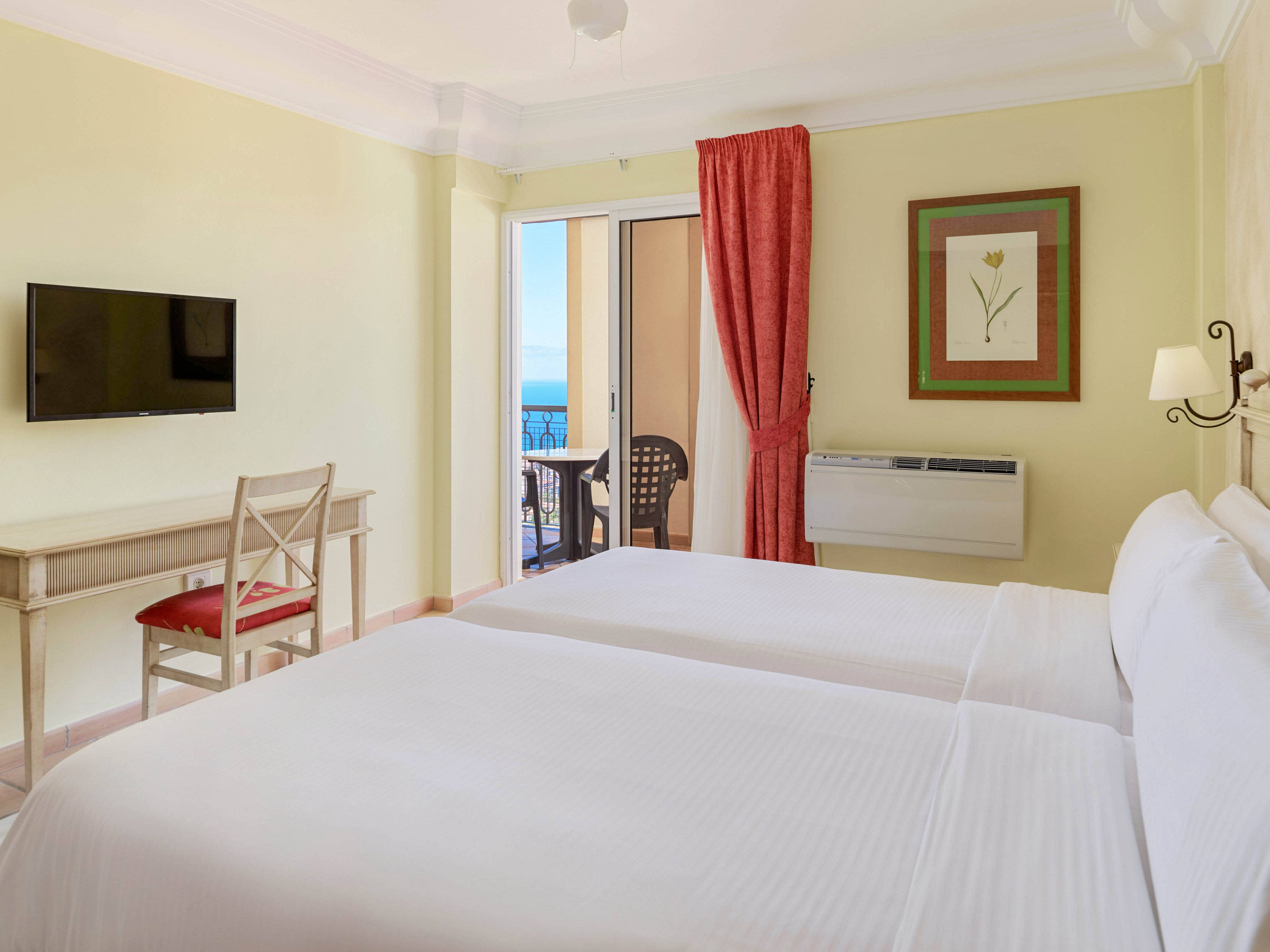 A tidy hotel room features two neatly made twin beds with white linens against soft yellow walls. A flat-screen television is mounted above a simple desk and chair with a red cushion. Red curtains frame a glass door that opens to a balcony with outdoor chairs and a distant ocean view. A wall-mounted air conditioning unit and a framed floral artwork complete the cozy, inviting interior.