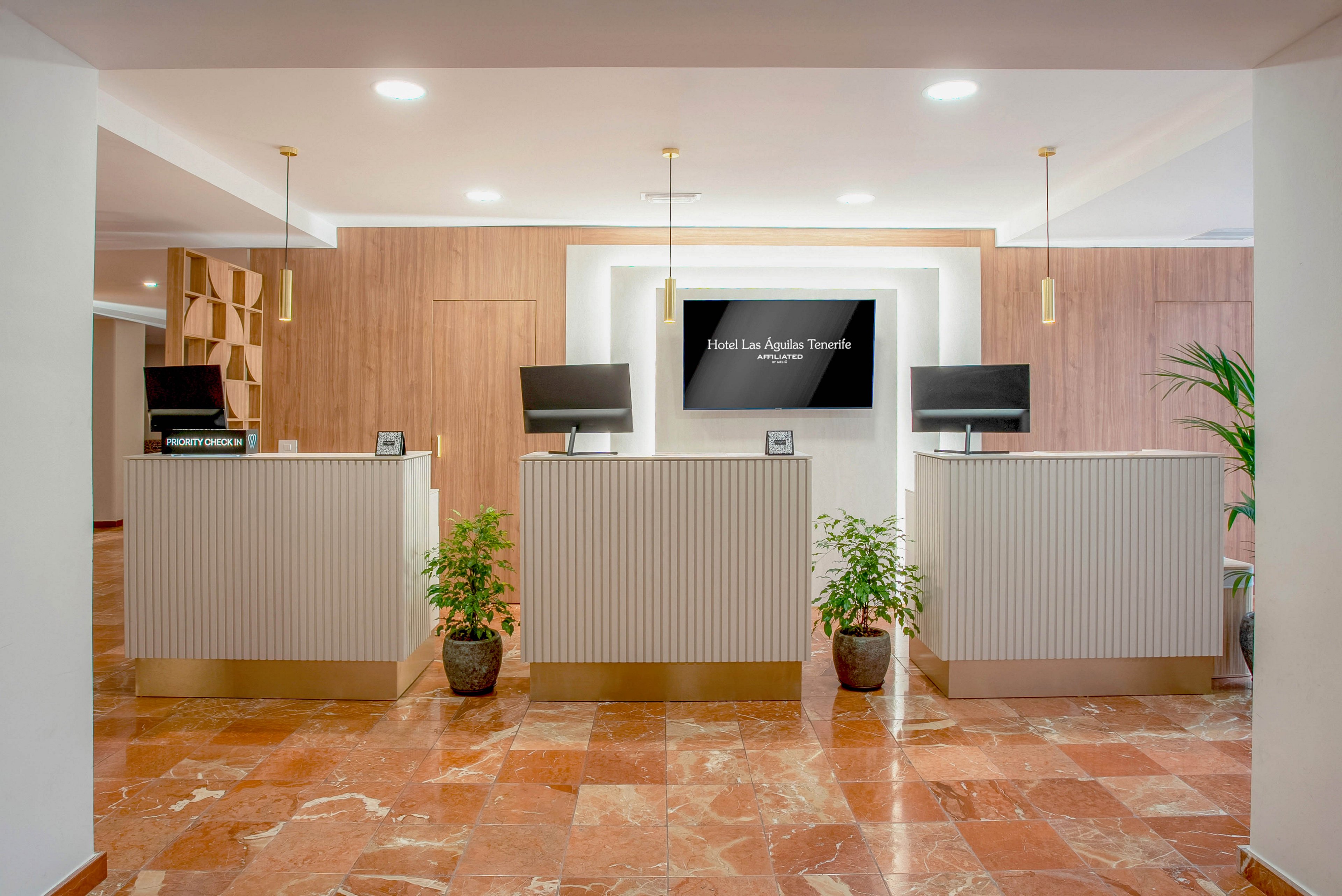 Contemporary hotel reception area featuring three front desks with computers and small potted plants. The space has warm wooden wall panels, a central digital screen displaying 'Hotel Las Águilas Tenerife', and pendant lights hanging from the ceiling. Marble-style floor tiles add texture and color to the clean, minimalist interior. A sign reading 'RECEPTION' is visible on one of the desks, emphasizing the check-in function of the space.