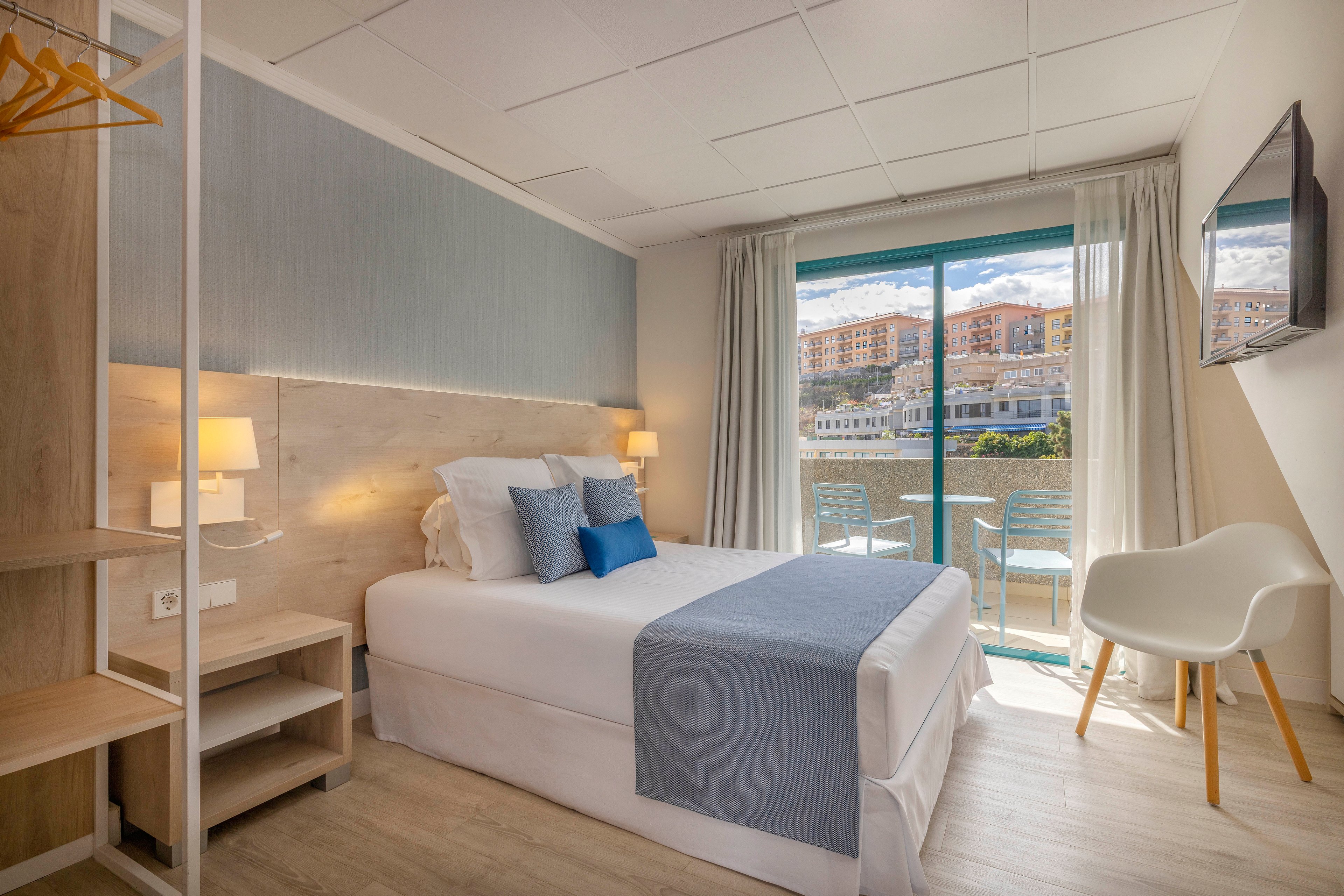 A neatly made double bed with white linens and blue accents is the focal point of a modern hotel room. Soft lighting from bedside lamps and natural light from a large glass door create a bright, welcoming atmosphere. Outside, a balcony with chairs overlooks residential buildings under a partly cloudy sky. Minimalist furniture, light wood flooring, and neutral tones emphasize a clean, contemporary design.