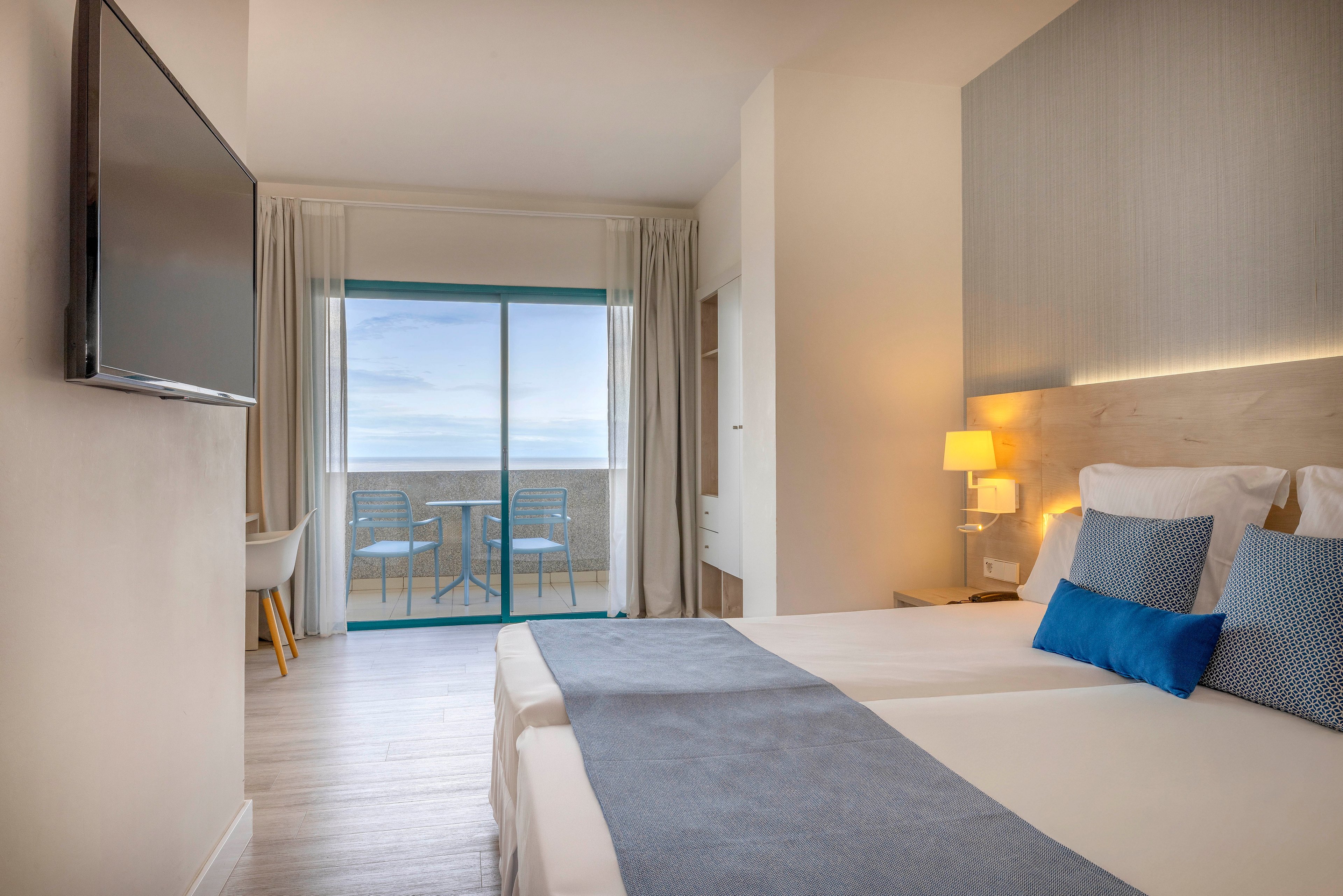 A contemporary hotel room features two neatly made twin beds with white linens and blue accent runners and pillows. A large flat-screen television is mounted on the wall opposite the beds, and a small bedside table holds a lit lamp. Floor-to-ceiling curtains frame a sliding glass door that opens to a balcony with two chairs and a table. Beyond the balcony, a calm ocean view is visible under a soft sky, creating a serene and inviting atmosphere.