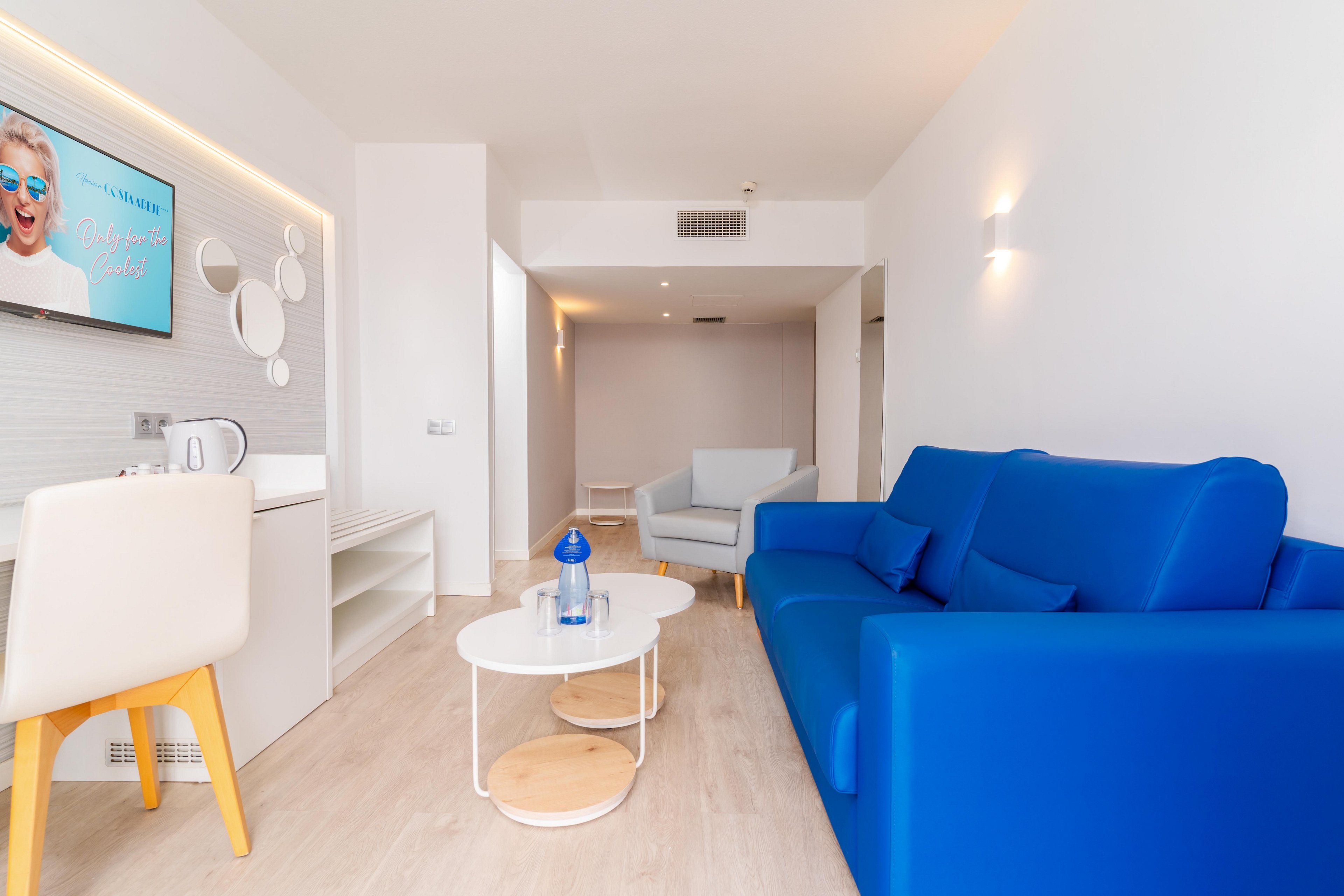 A contemporary living room featuring a vibrant blue sofa and minimalist decor. The space includes a white coffee table with a water bottle, a kitchenette, and a wall-mounted TV displaying a smiling woman with text 'Enjoy your stay'. The room is well-lit with natural and artificial lighting, creating a bright and inviting atmosphere.