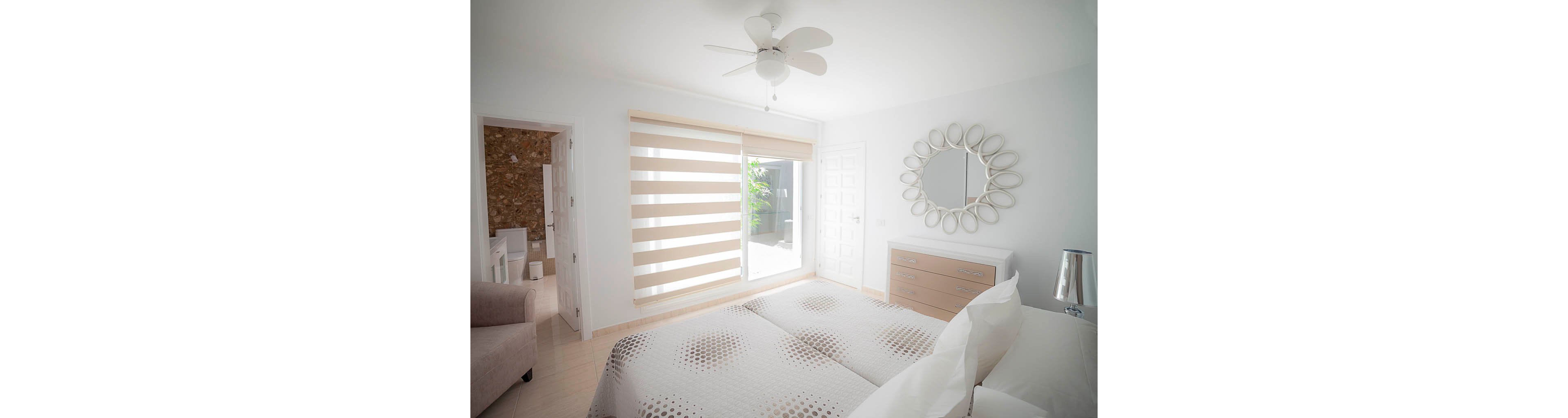 A bright, minimalist bedroom features a neatly made double bed with patterned bedding. Large horizontal blinds cover a sliding glass door that opens to an outdoor area with greenery. A round decorative mirror hangs above a light-colored dresser, and a ceiling fan with a light fixture is centered above the room. Soft neutral tones and natural light create an airy, tranquil atmosphere.