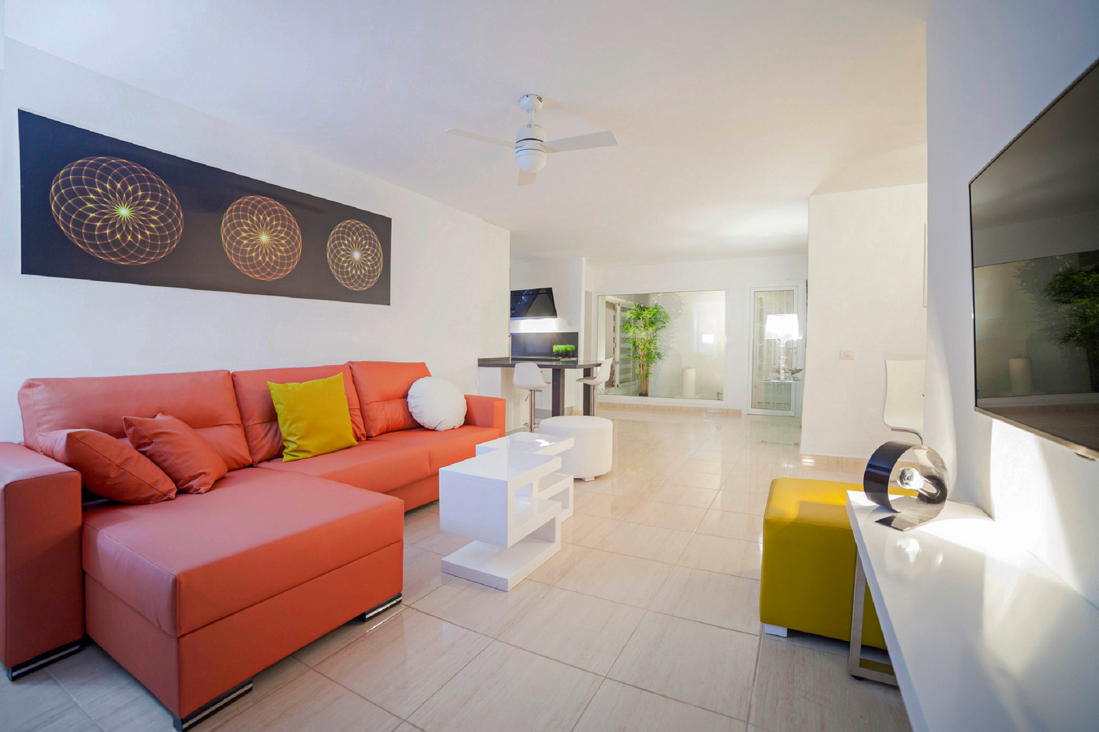 Contemporary living room interior featuring a large coral sectional sofa with colorful cushions. The space is decorated in a minimalist style with white walls, tiled flooring, and sleek modern furniture. A wall-mounted television, ceiling fan, and abstract artwork add visual interest. Soft lighting and neutral tones create an airy, inviting atmosphere.