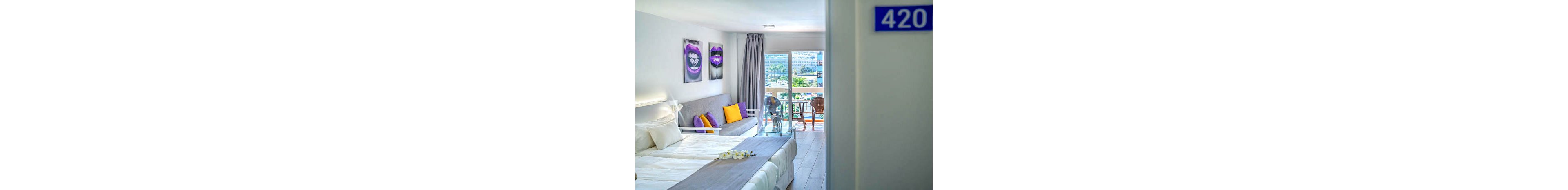 A bright, contemporary hotel room features a neatly made bed, a gray sofa with colorful pillows, and modern wall art. The room opens to a balcony with two chairs and a table, overlooking a cityscape with palm trees. The door displays the number '420', and the interior is decorated in a cool, minimalist palette with pops of yellow and purple.