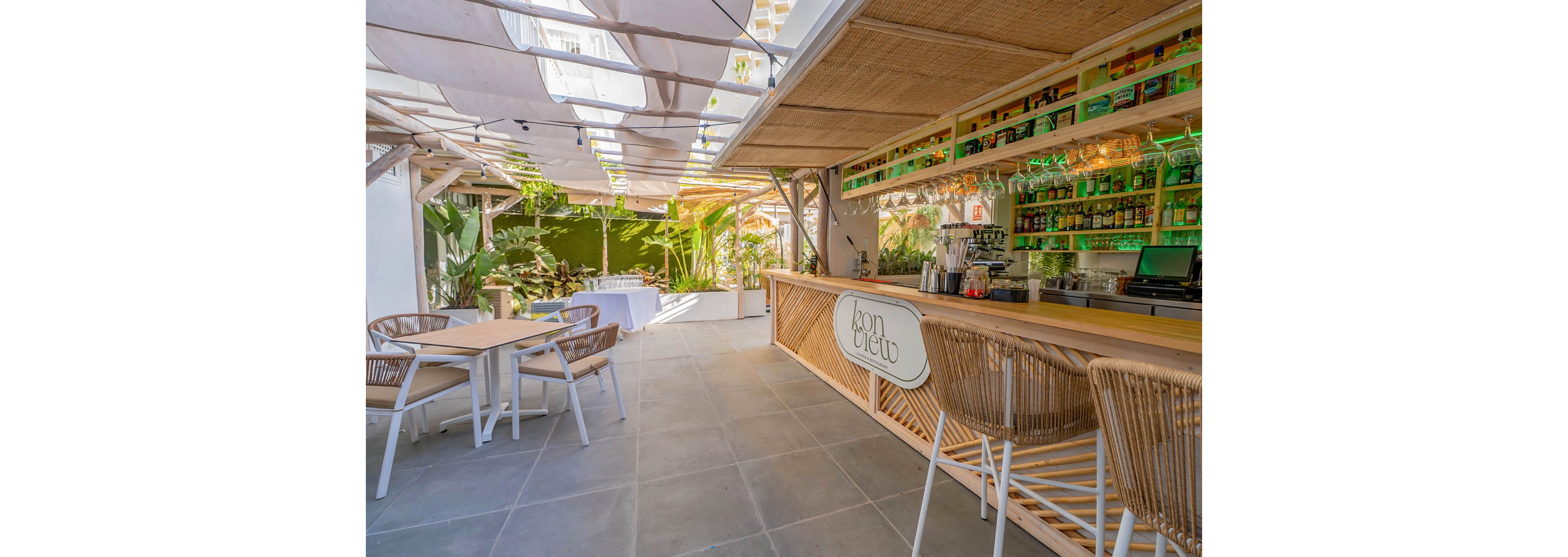 A stylish outdoor bar features a long wooden counter with wicker bar stools and a matching table set. The setting is bright and airy, with natural light streaming through a partially covered roof and lush green plants in the background. Shelves behind the bar display various bottles, illuminated by green accent lighting. The overall mood is relaxed and inviting, ideal for social gatherings or casual dining. The sign on the bar reads 'abon bistro'.