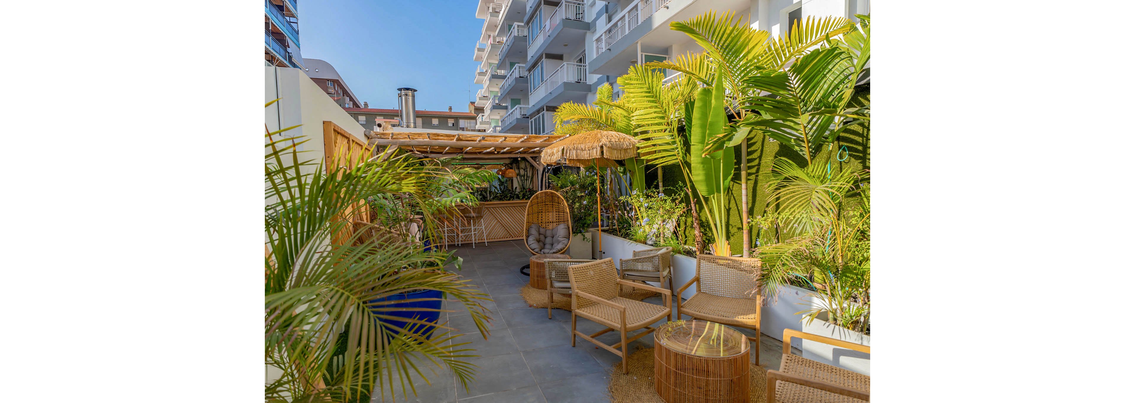The image shows a modern outdoor lounge area featuring wicker chairs, tables, and a hanging egg chair. Lush tropical plants line the space, creating a relaxing atmosphere. The setting is adjacent to a contemporary apartment building, with sunlight highlighting the greenery and seating. The mood is casual and inviting, with a natural palette of greens and tans.