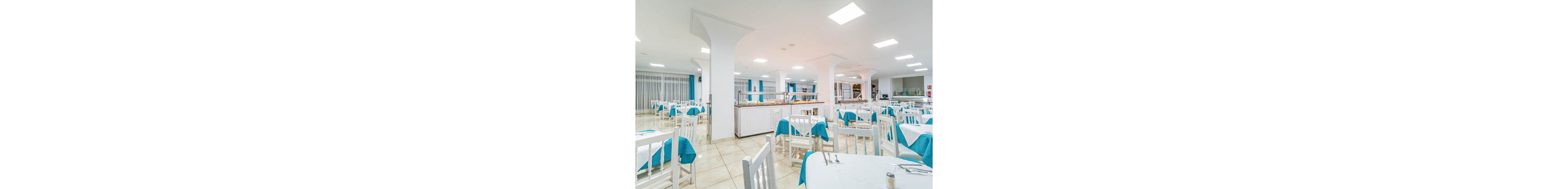 A spacious, well-lit restaurant dining area features white tables and chairs with turquoise tablecloth accents. The setting includes a central buffet station and large windows with sheer curtains, creating an inviting and clean atmosphere. The room is empty, emphasizing the organized table settings and contemporary decor. Overhead lighting and tiled floors contribute to the fresh, modern style.