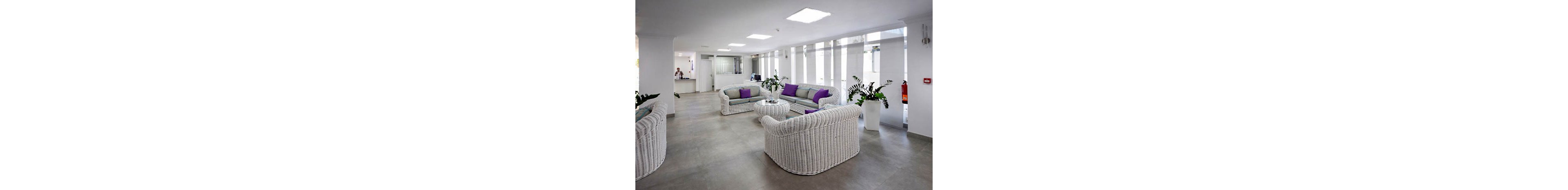 The image shows a spacious waiting area furnished with white wicker sofas and glass tables. Purple cushions add a pop of color to the neutral palette, while large potted plants provide greenery. The setting appears clean and contemporary, with bright overhead lighting and a reception desk visible in the background. No visible text or numbers are present in the scene.