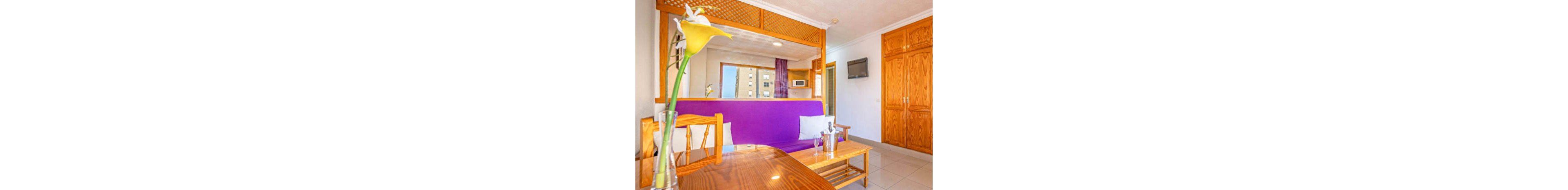 A modern hotel room features a vibrant purple sofa with white pillows, a wooden table, and a yellow calla lily in a glass vase. The setting includes wooden furniture, a mounted TV, and a window view of a city building. The mood is cheerful and welcoming, with warm wood tones and natural light enhancing the space. A metal ice bucket and two glasses are placed on the coffee table, suggesting relaxation or celebration.