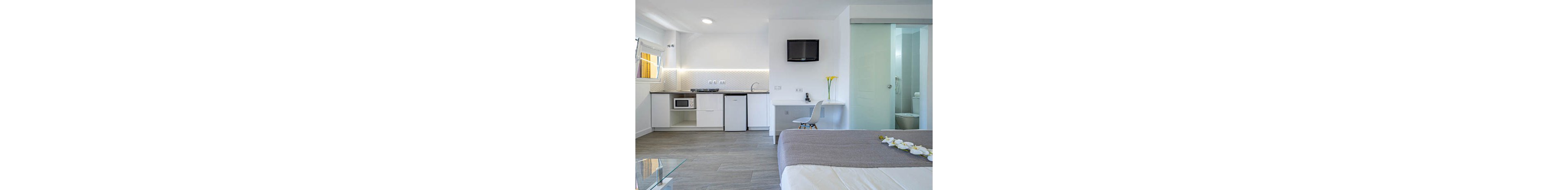 A contemporary studio apartment features a compact kitchenette with a microwave, mini fridge, and sink. The space includes a small desk with a chair, a wall-mounted television, and a glass sliding door leading to a bathroom. The room is decorated in neutral tones with bright natural light and minimalist style, and a bed with a gray blanket and white flowers is visible in the foreground.