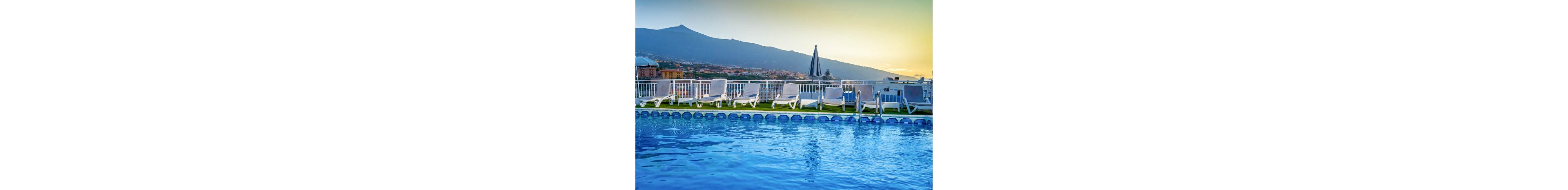 A serene outdoor swimming pool is featured in the foreground, surrounded by white lounge chairs and a closed umbrella. The setting overlooks a scenic mountain and cityscape, with the sky transitioning from blue to warm hues as the sun sets. The water in the pool reflects the tranquil atmosphere, and the area appears empty and peaceful.