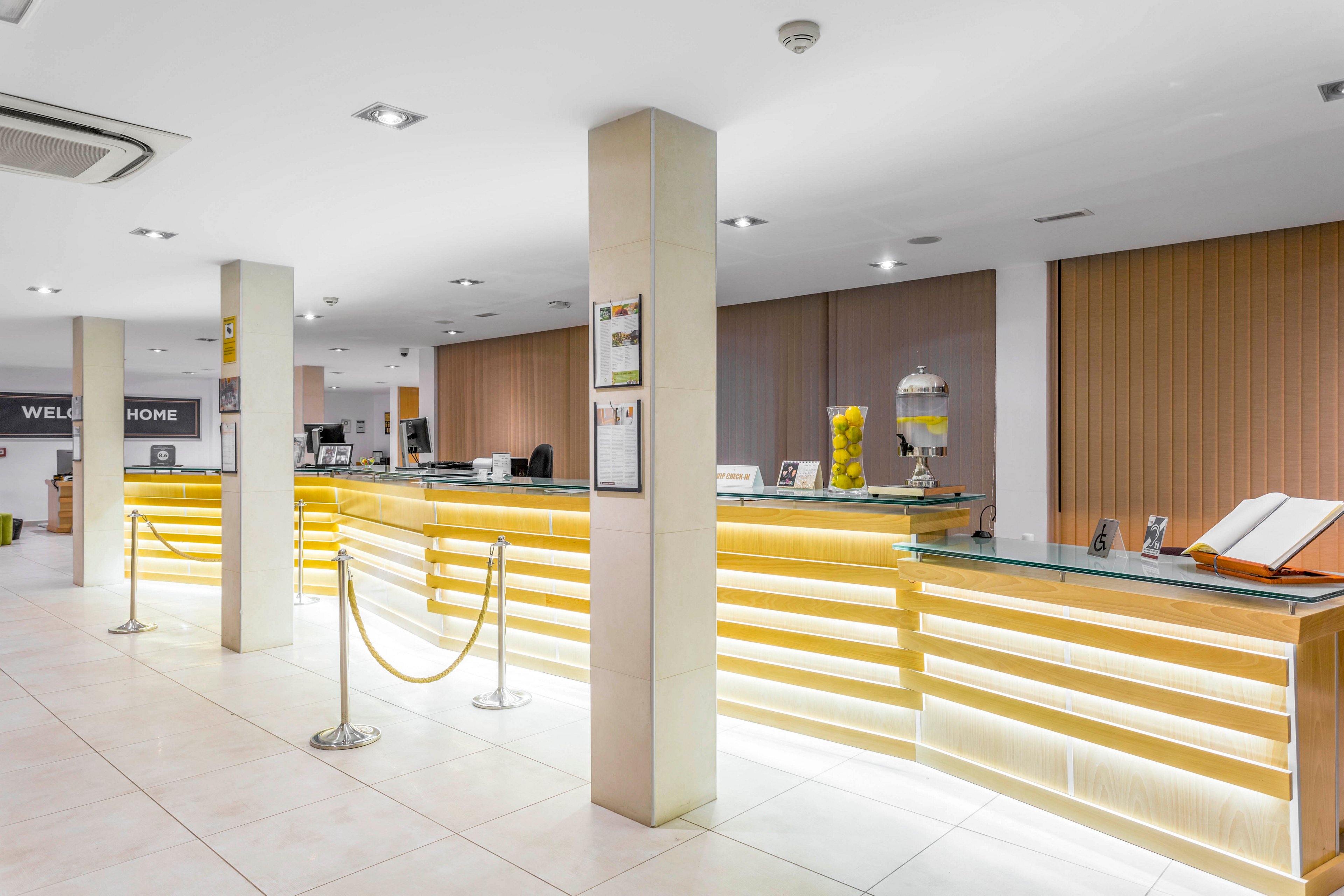 A spacious hotel reception area features a long, illuminated front desk with glass countertops. The setting is clean and contemporary, with tiled floors, vertical blinds, and ceiling spotlights. A rope barrier guides guests toward the check-in area, where computers and office equipment are visible. Decorative elements such as framed notices, a glass drink dispenser, and an open guest book add detail to the bright, welcoming space.