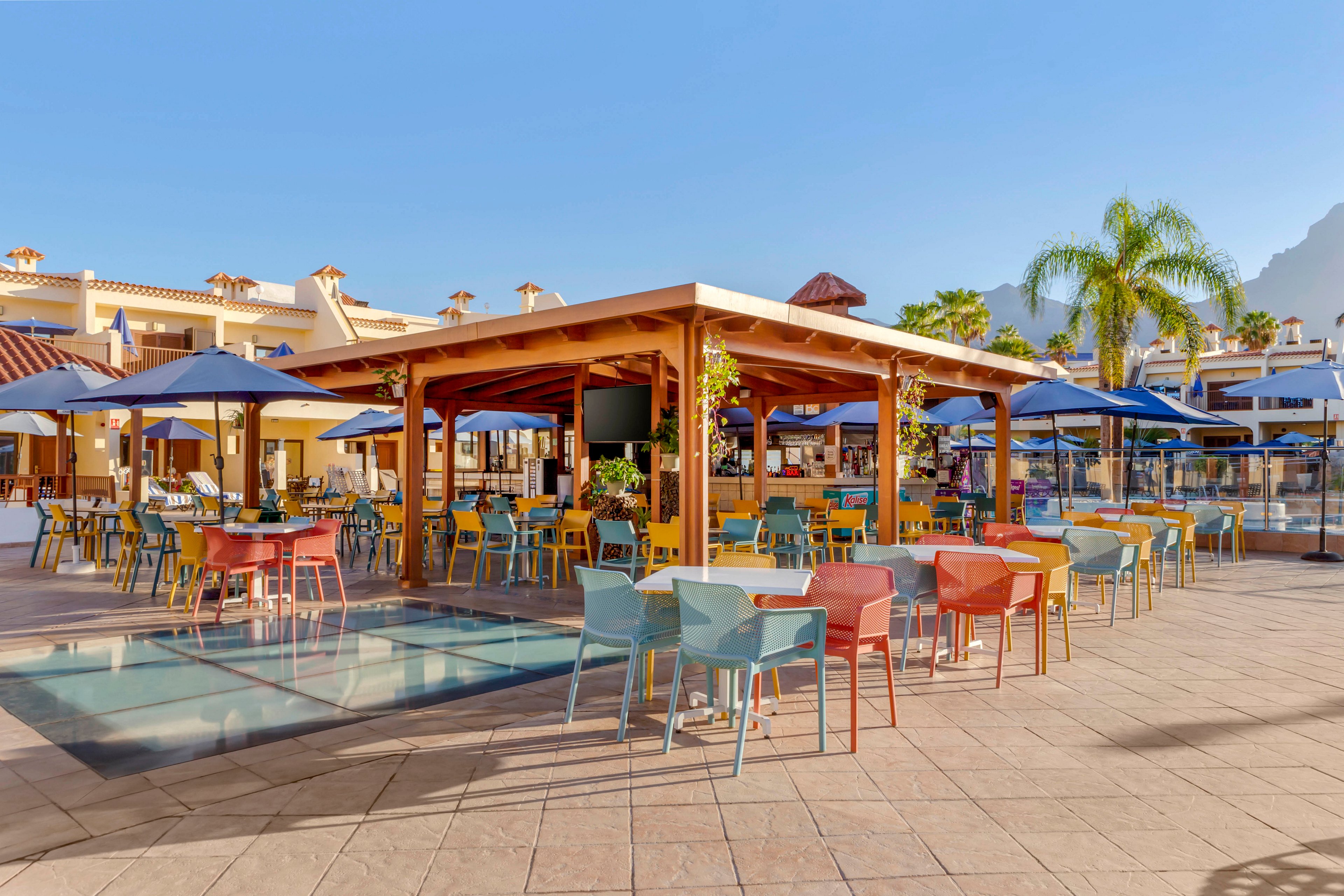An open-air bar with a wooden pergola stands at the center of a resort patio. Colorful chairs and tables are arranged around the bar, many shaded by large blue umbrellas. The scene is bright and sunny, with clear blue sky and palm trees adding to the vacation atmosphere. Surrounding buildings with tiled roofs and distant mountains complete the relaxed holiday setting.