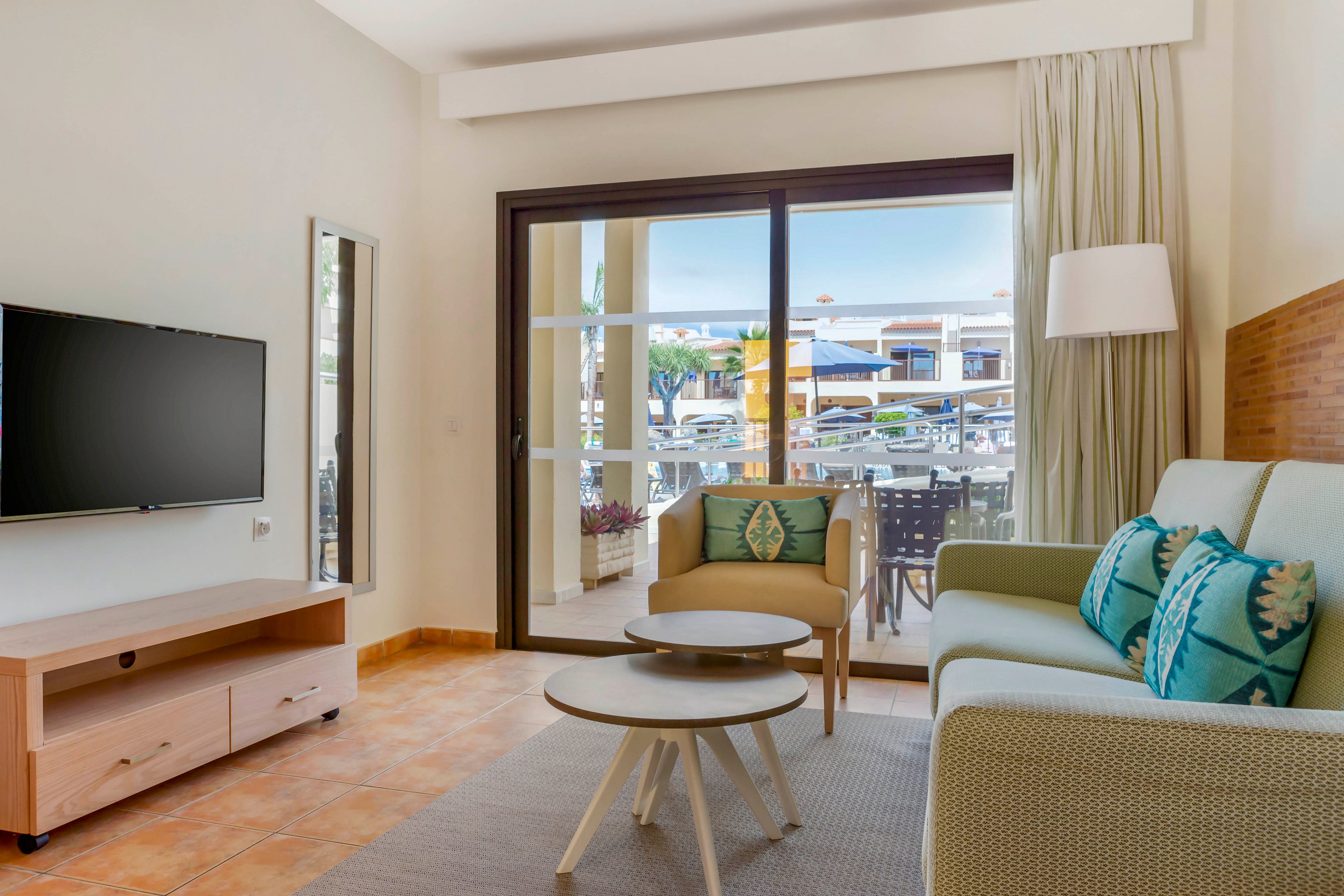 A modern living room features a light green sofa, armchair, and nested coffee tables. Large sliding glass doors open to an outdoor terrace with patio furniture and views of neighboring buildings. A flat-screen television is mounted above a wooden media console on the left wall. Neutral walls, tiled flooring, and soft natural light create an airy, welcoming atmosphere.