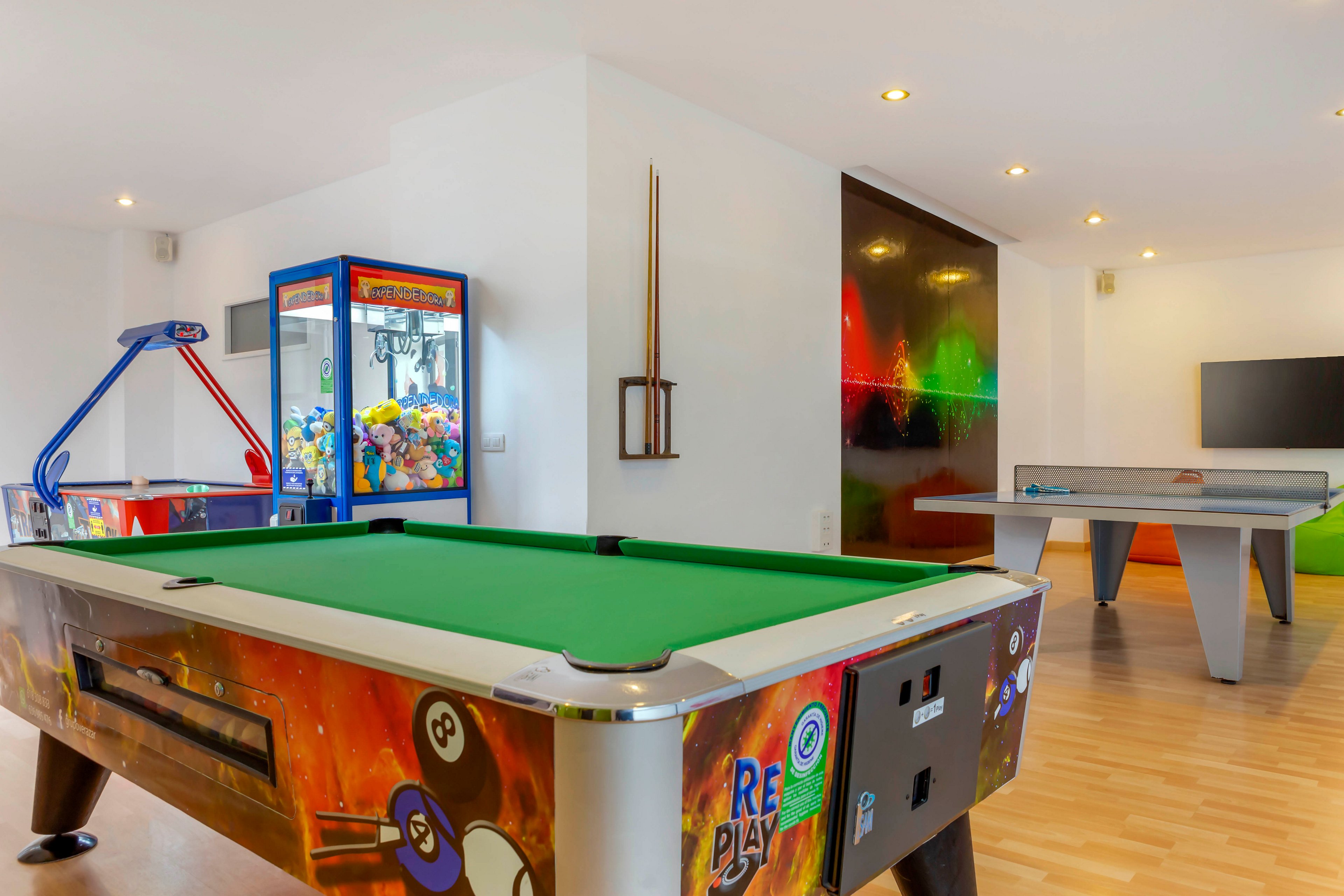A contemporary game room features a green-felt pool table in the foreground. In the background, there is an air hockey table and a claw crane machine filled with colorful plush toys. The room has light wooden flooring, white walls, and recessed ceiling lights creating a bright atmosphere. A sleek wall-mounted television and a vibrant abstract wall panel add to the modern entertainment setting.