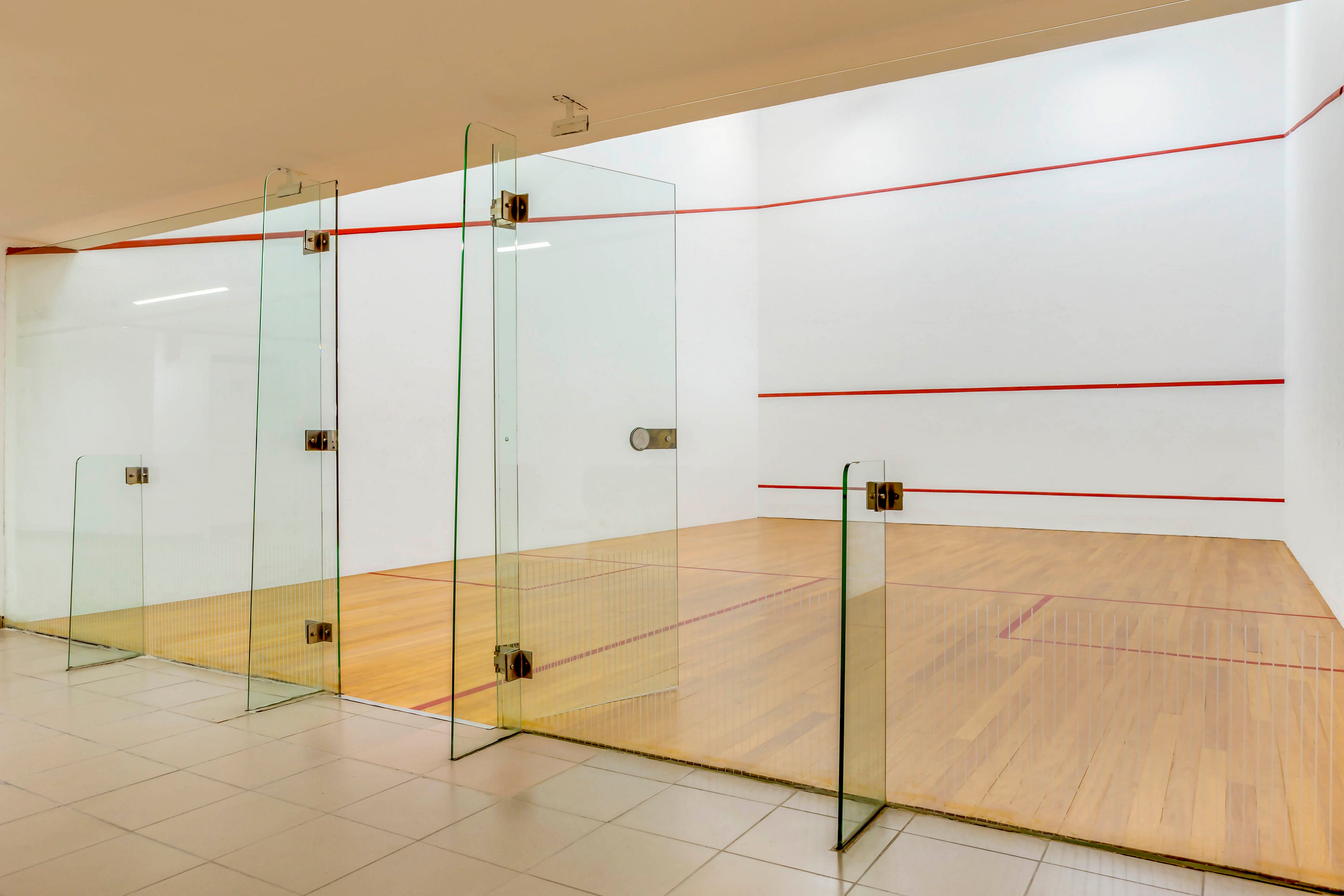 An indoor squash court is shown with clear glass walls and doors opening onto a tiled corridor. The court features a polished wooden floor and bright white walls marked with red boundary lines. Overhead lighting illuminates the clean, modern sports facility. The space is empty, emphasizing the geometry of the court and the transparency of the glass panels.