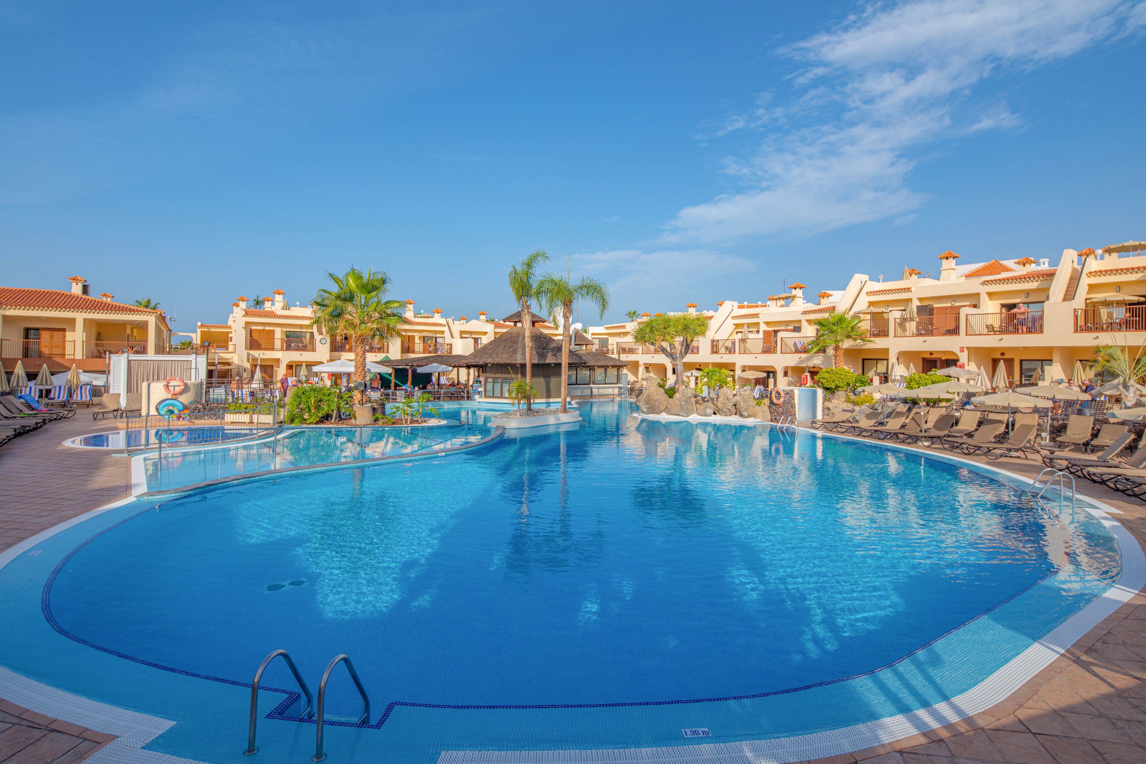 A large curved swimming pool sits at the center of a sunny resort complex. Multi-story vacation apartments with balconies surround the pool area. Numerous lounge chairs and umbrellas line the tiled pool deck, with palm trees adding a tropical touch. The clear blue sky and calm water create a relaxed holiday atmosphere.