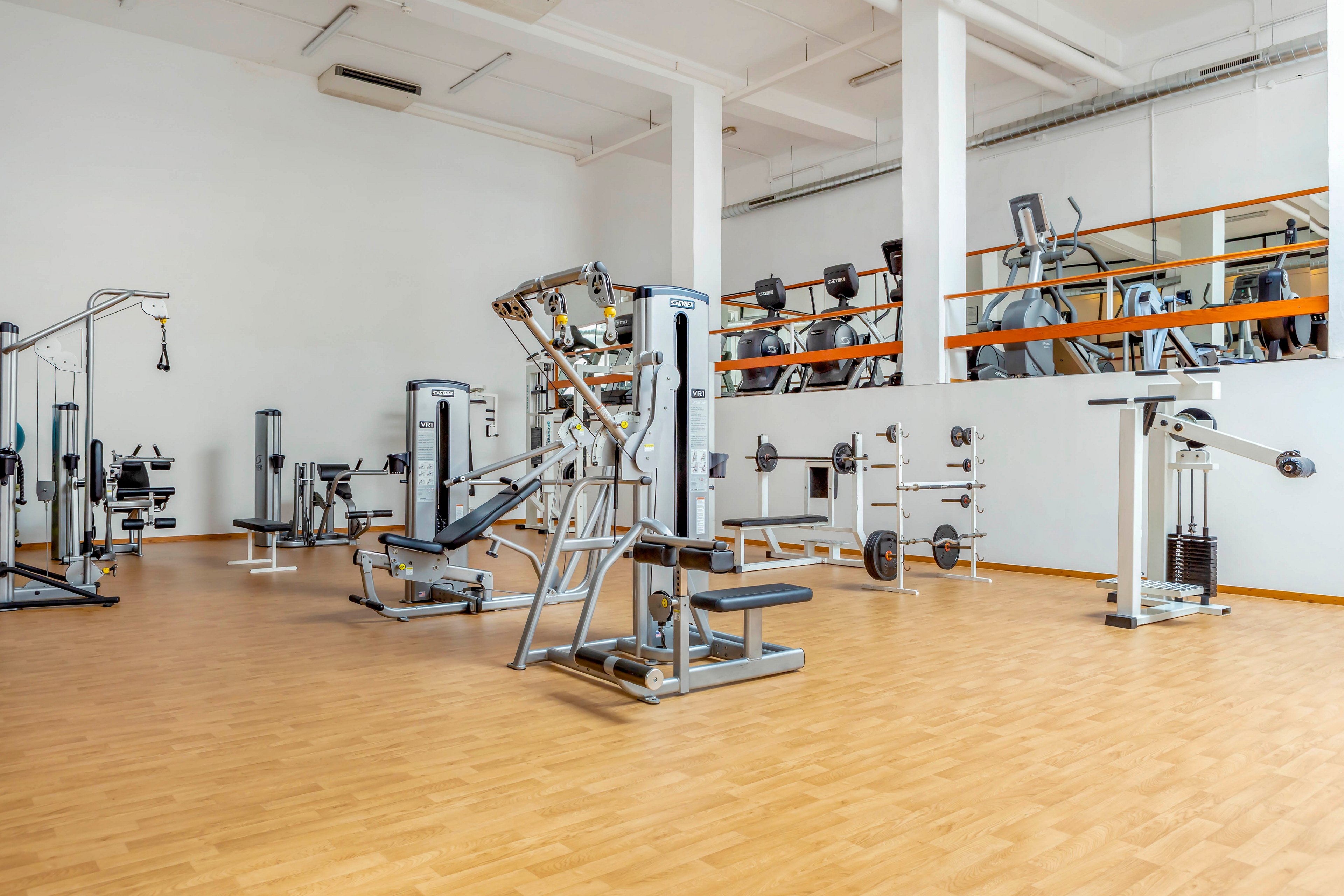 A spacious, modern gym interior features various strength training machines and free weights. The room has light-colored wooden flooring and white walls, creating a clean and bright atmosphere. Multiple weight machines, benches, and cable systems are neatly arranged across the open floor. In the background, additional equipment is visible on a raised mezzanine level behind an orange railing.