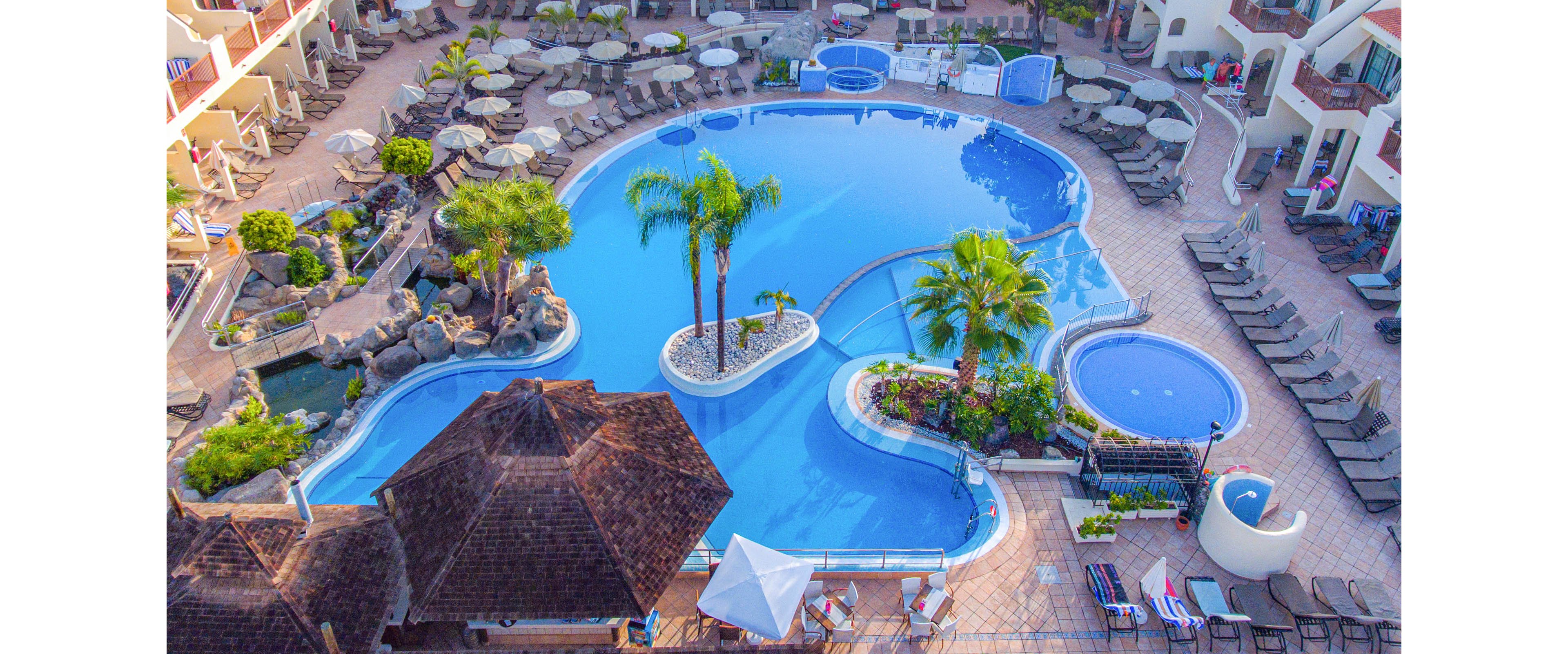 A large resort swimming pool complex is captured from an aerial perspective. The main freeform pool is surrounded by sun loungers, umbrellas, and several smaller circular pools. Palm trees and landscaped islands decorate the center of the pool, creating a tropical atmosphere. Buildings and terraces encircle the pool area, emphasizing a vacation and leisure setting.