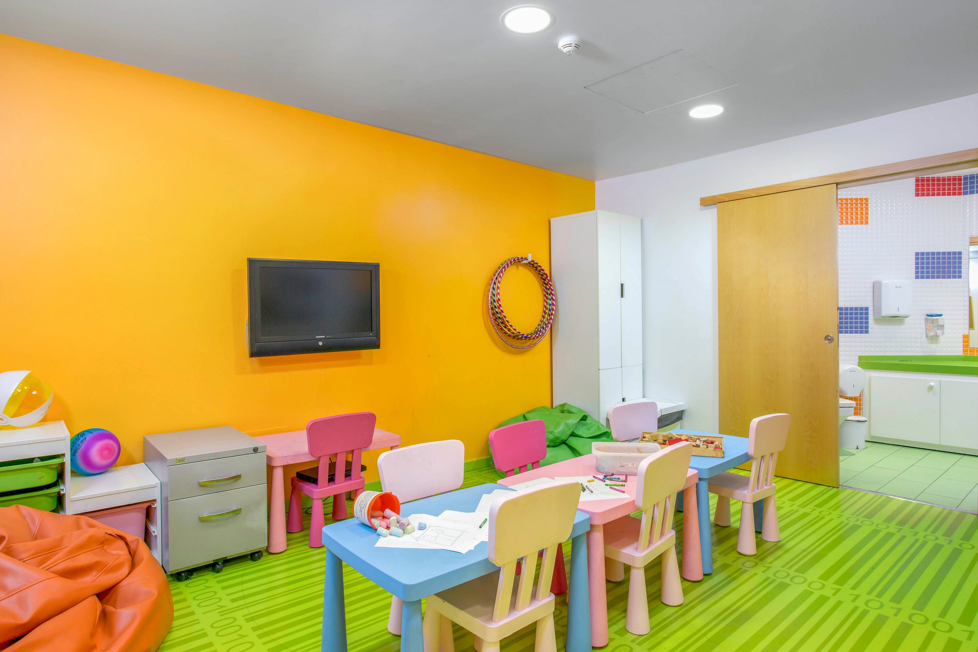 Brightly colored children's playroom featuring small tables and chairs arranged for group activities. The room has an orange accent wall with a mounted flat-screen television and playful decor. Green striped flooring and pastel furniture create a cheerful, inviting atmosphere. In the background, storage cabinets and a partially open sliding door reveal an adjoining area with additional colorful details.