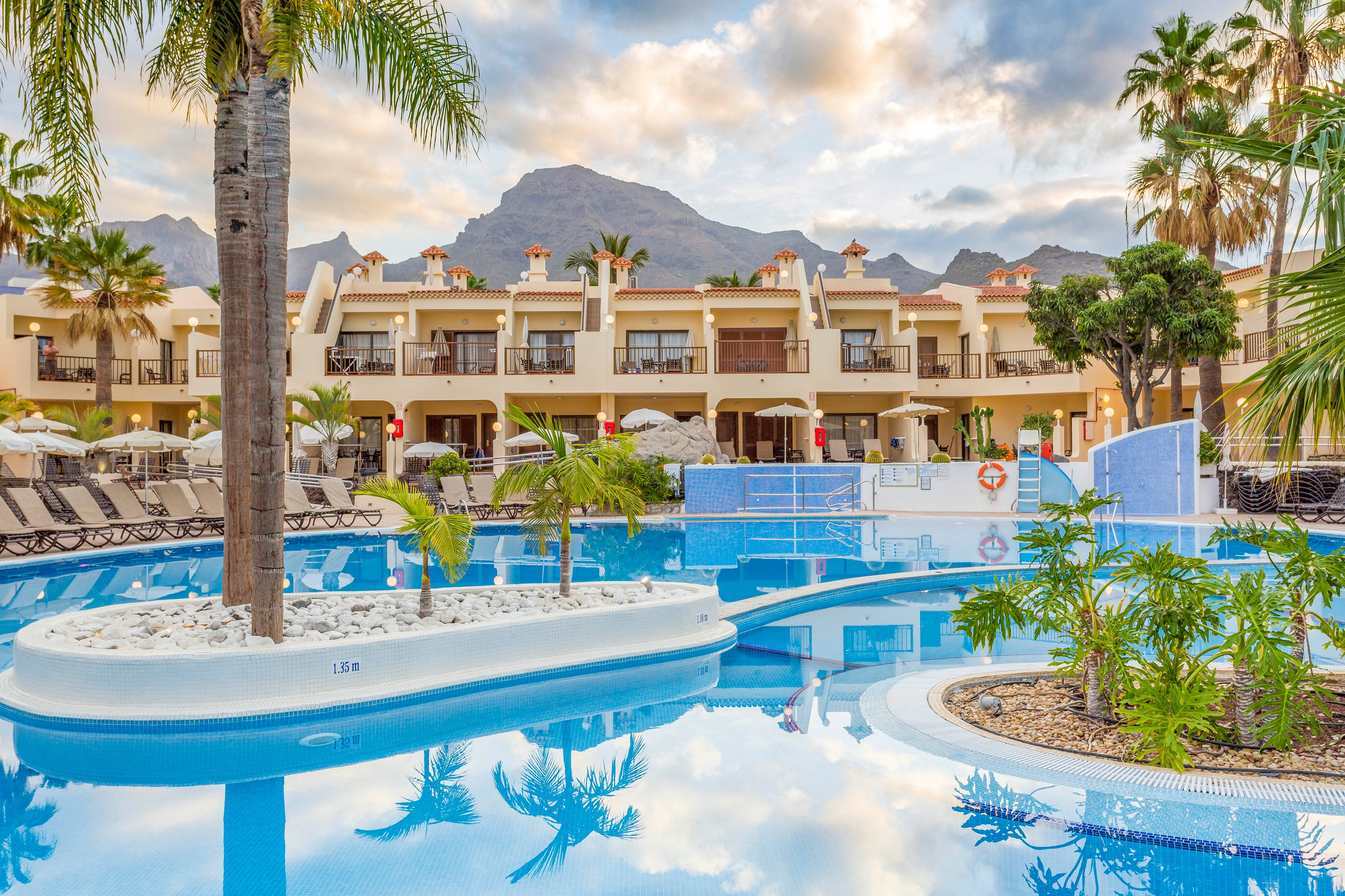 A large resort swimming pool reflects palm trees and surrounding buildings under a soft evening sky. Sun loungers and parasols line the poolside terraces of the multi-story accommodation. Mountains rise in the background, adding a dramatic natural backdrop to the tranquil scene. The calm blue water and warm architecture create a relaxing vacation atmosphere.