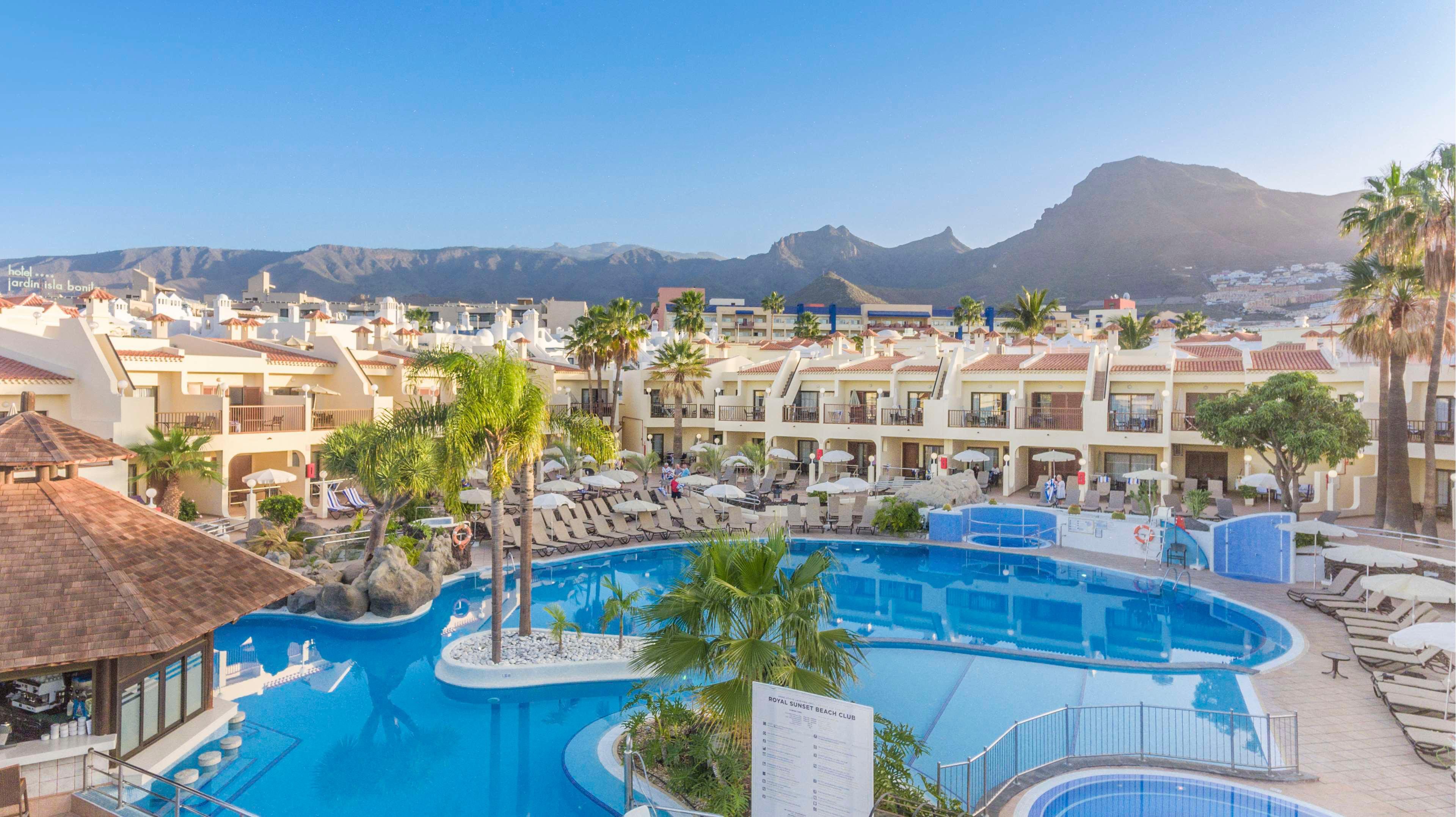 A large outdoor swimming pool curves through a sunny resort complex lined with lounge chairs and umbrellas. Multi-story vacation apartments surround the pool area, creating a relaxed holiday atmosphere. Palm trees, a pool bar structure, and clear blue water emphasize the tropical feel. Distant mountains rise under a bright blue sky, framing the leisure scene.