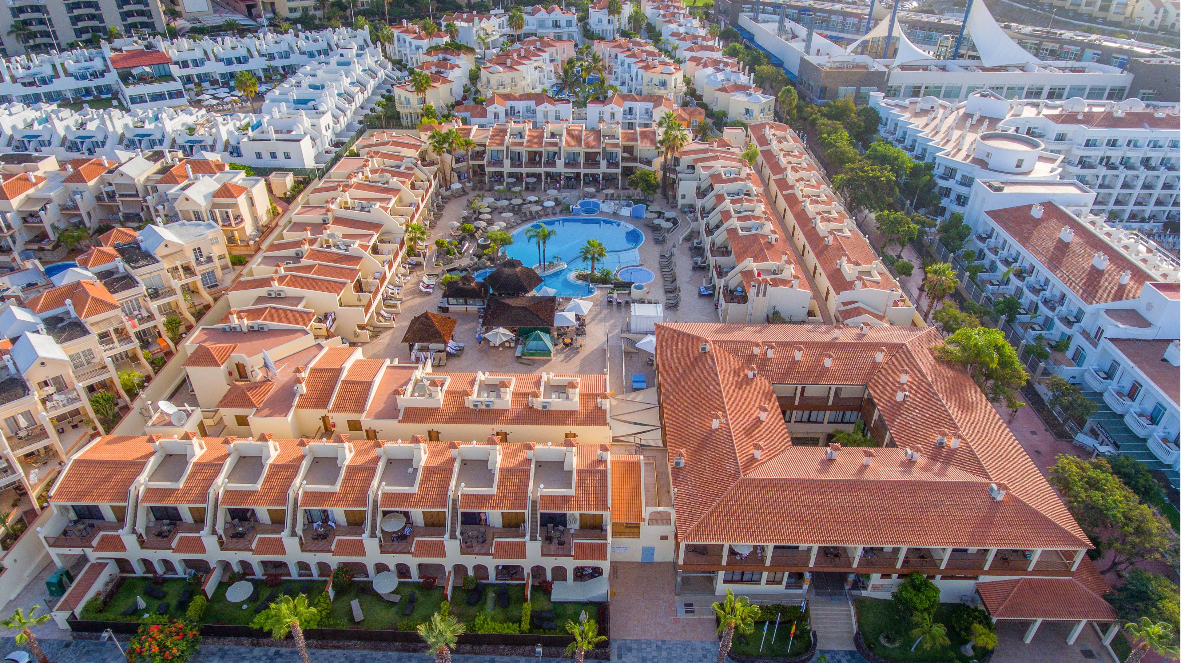 A large resort complex with terracotta roofs is captured from an aerial perspective. The buildings surround a central circular swimming pool with sun loungers and umbrellas. Palm trees and landscaped gardens are visible throughout the property, adding greenery between the structures. Nearby streets and additional white buildings indicate an urban coastal or tourist destination setting.
