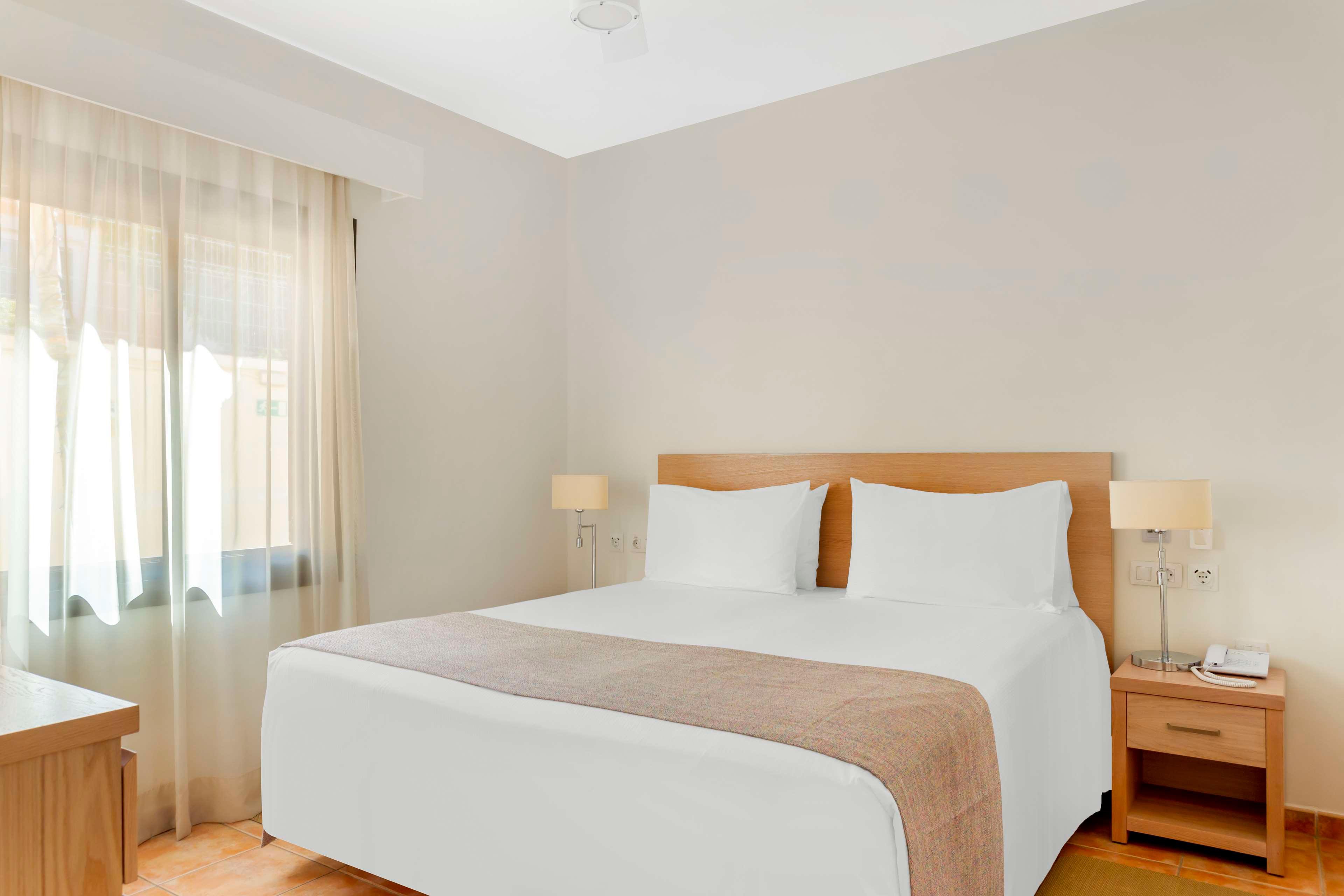 A neatly made double bed with white linens and a beige throw is centered in a bright hotel room. Natural light filters through sheer curtains covering a large window on the left. Matching wooden nightstands with modern lamps flank the bed, and a small desk is visible in the corner. The room features neutral walls and warm-toned flooring, creating a clean and inviting atmosphere.