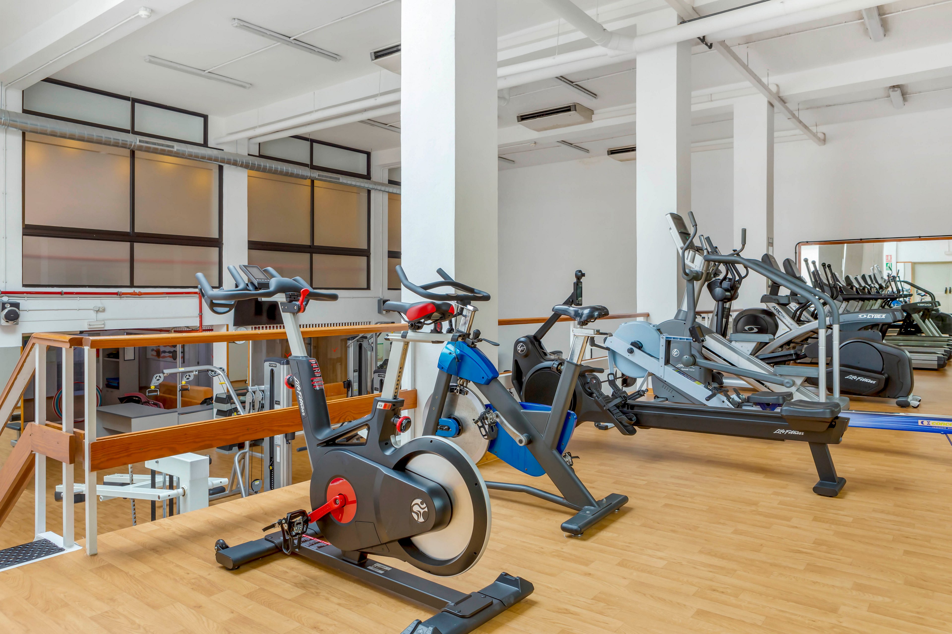 A bright, modern gym interior features several pieces of cardio equipment, including stationary bikes and elliptical machines. The space has light wood flooring and white walls, creating a clean and spacious atmosphere. Large windows and glass panels allow natural light to fill the room. A staircase with wooden handrails leads to another level of the fitness area.