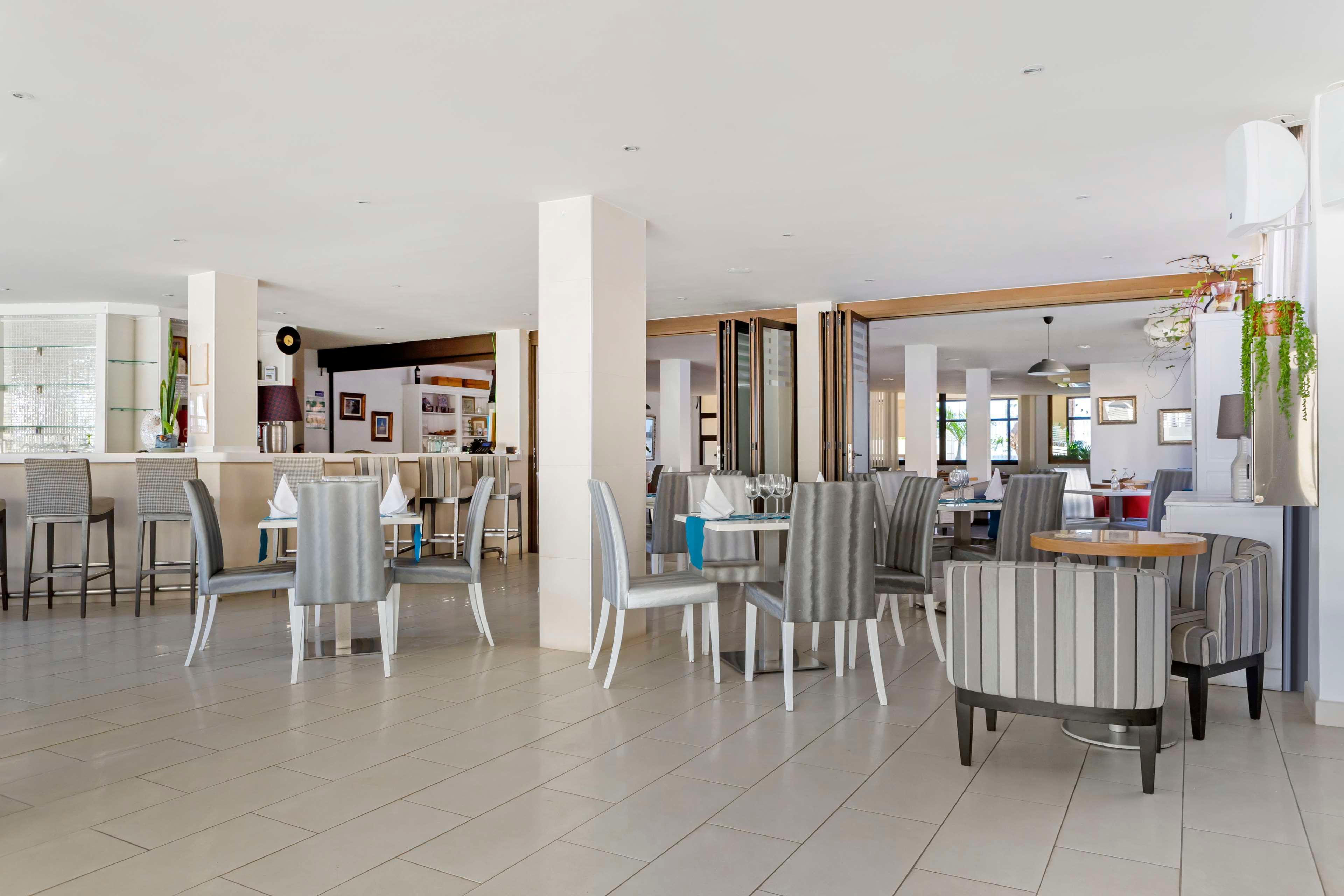 Spacious, contemporary restaurant interior with neatly arranged tables and chairs. The room features light tiled flooring, white walls, and large windows that allow natural daylight to fill the space. A bar area with high stools is visible in the background, along with framed artwork on the walls. Neutral grey and white tones dominate the decor, creating a clean and inviting atmosphere suitable for hotel guests or casual dining.