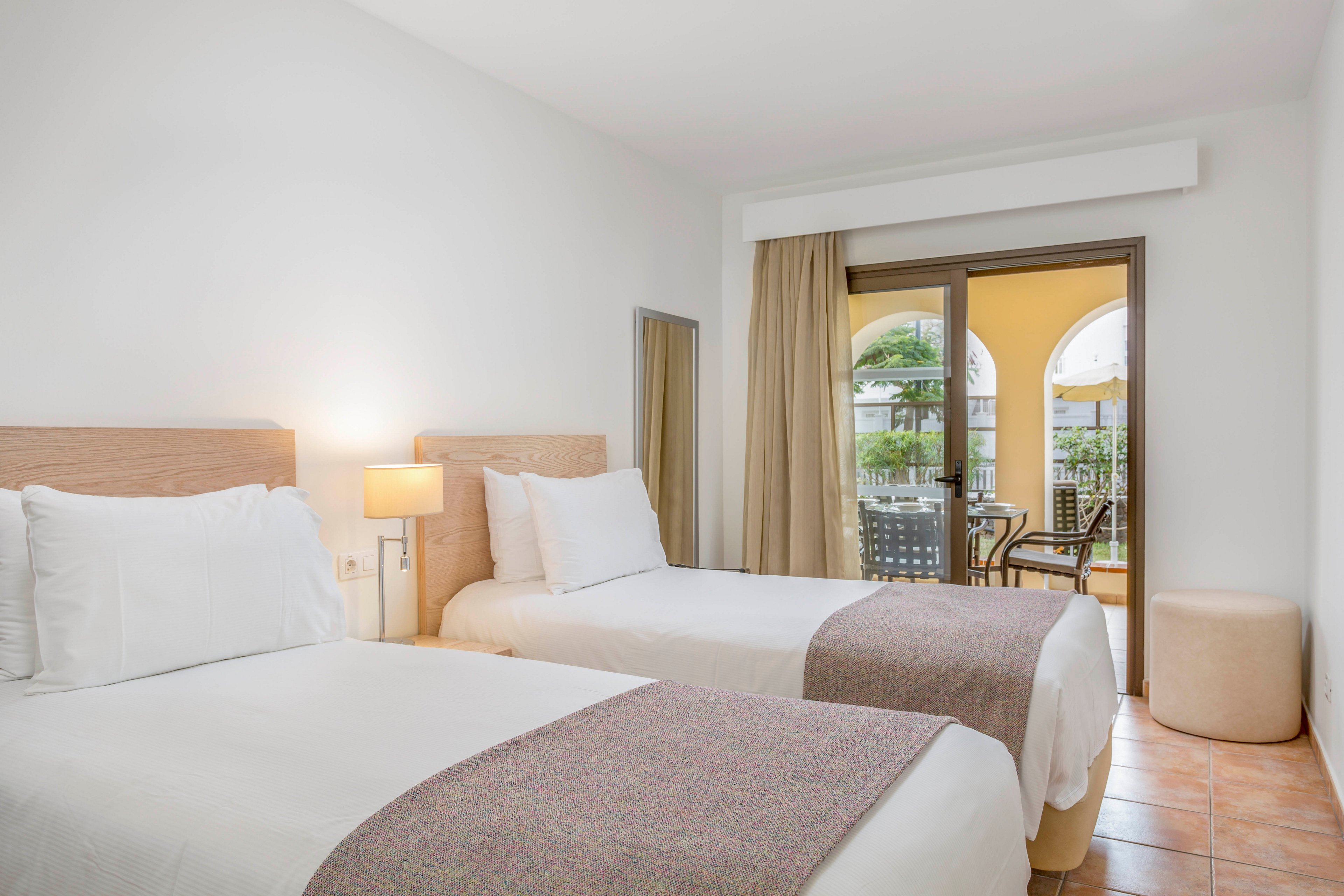A clean, modern hotel room features two neatly made twin beds with white linens and soft neutral throws. A bedside table with a warm-lit lamp sits between the beds, adding a cozy touch to the minimalist decor. Large glass doors open to a balcony with outdoor chairs and a view of arches and greenery. The room has tiled floors, light-colored walls, and simple furnishings that create an airy, inviting atmosphere.