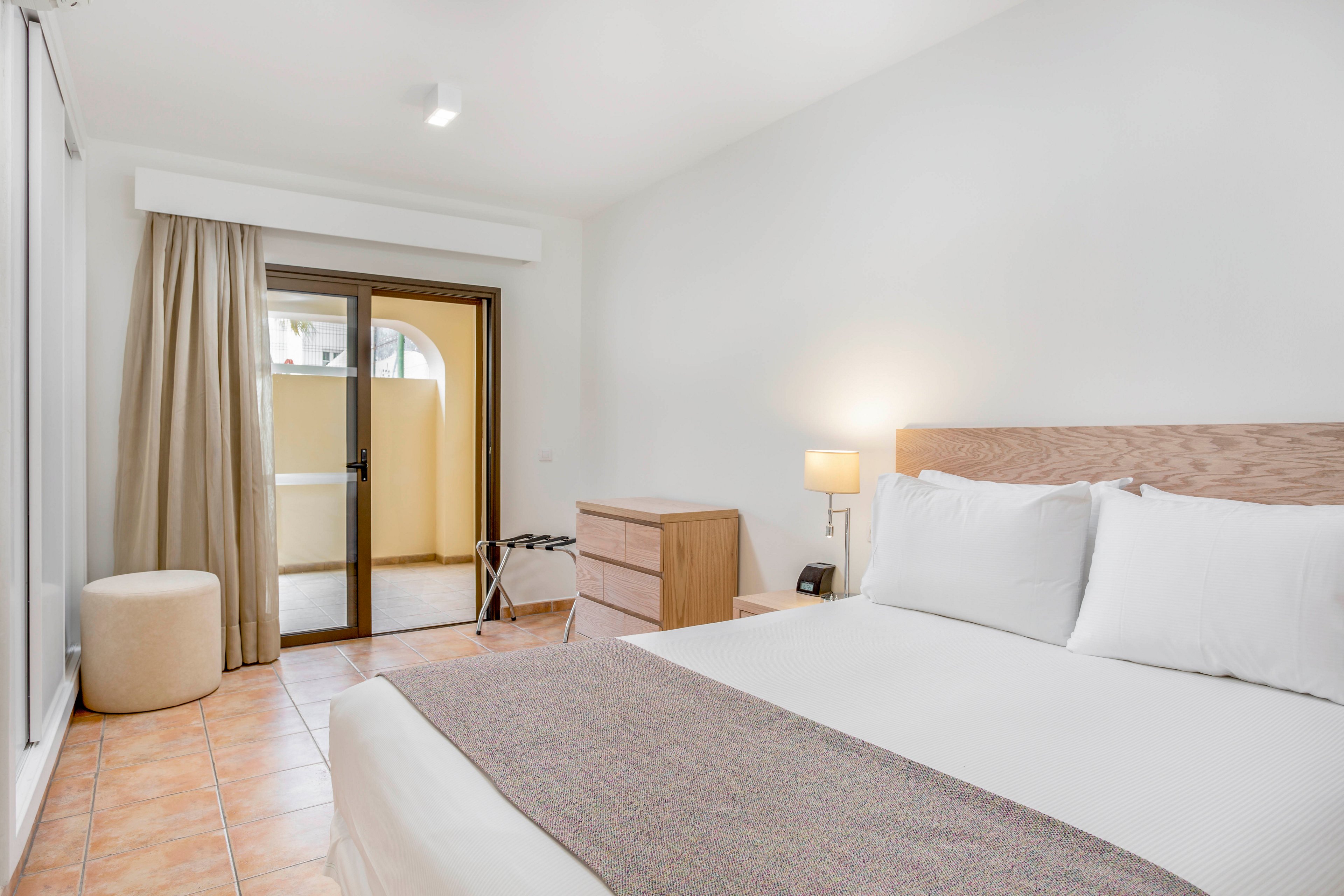 A bright, minimalist bedroom features a neatly made double bed with white linens and a neutral throw. The room includes a light wood nightstand with a lit table lamp and a small dresser. Sliding glass doors with beige curtains open to a private terrace area with tiled flooring. The overall design is clean and modern, emphasizing soft neutral tones and natural light.