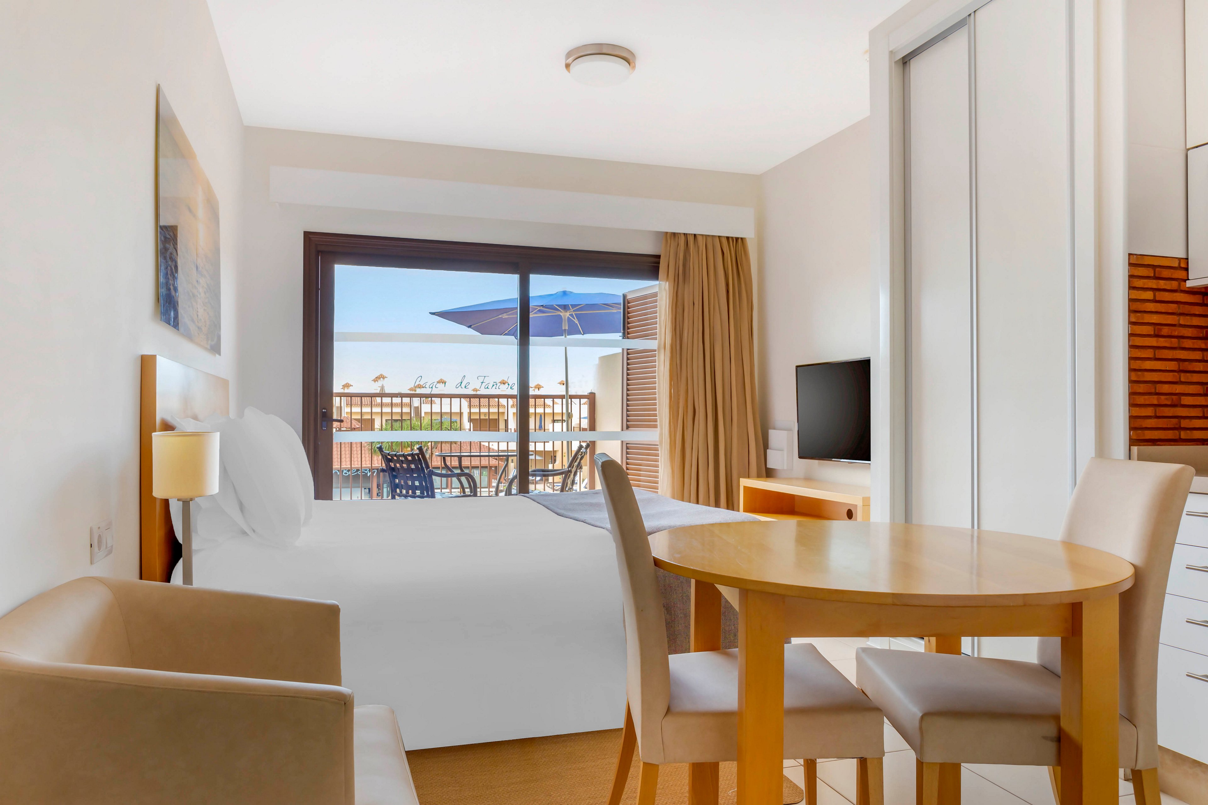 A modern hotel room features a neatly made double bed, small dining table, and compact seating area. Large sliding glass doors open to a balcony with outdoor chairs and a blue umbrella. Neutral tones and light wood furniture create a clean, minimalist atmosphere. A flat-screen television and built-in storage complete the cozy vacation space.